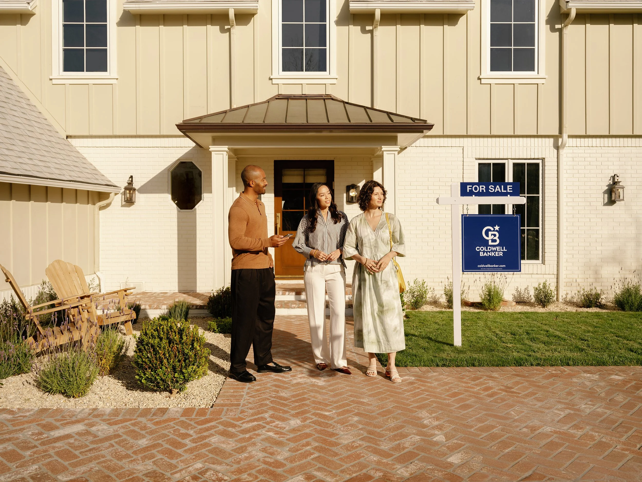 Three people standing in front of a house with a 'For Sale' sign, talking and smiling. The house has a light-colored exterior, multiple windows, and a small porch. The foreground shows a brick pathway and some landscaping.