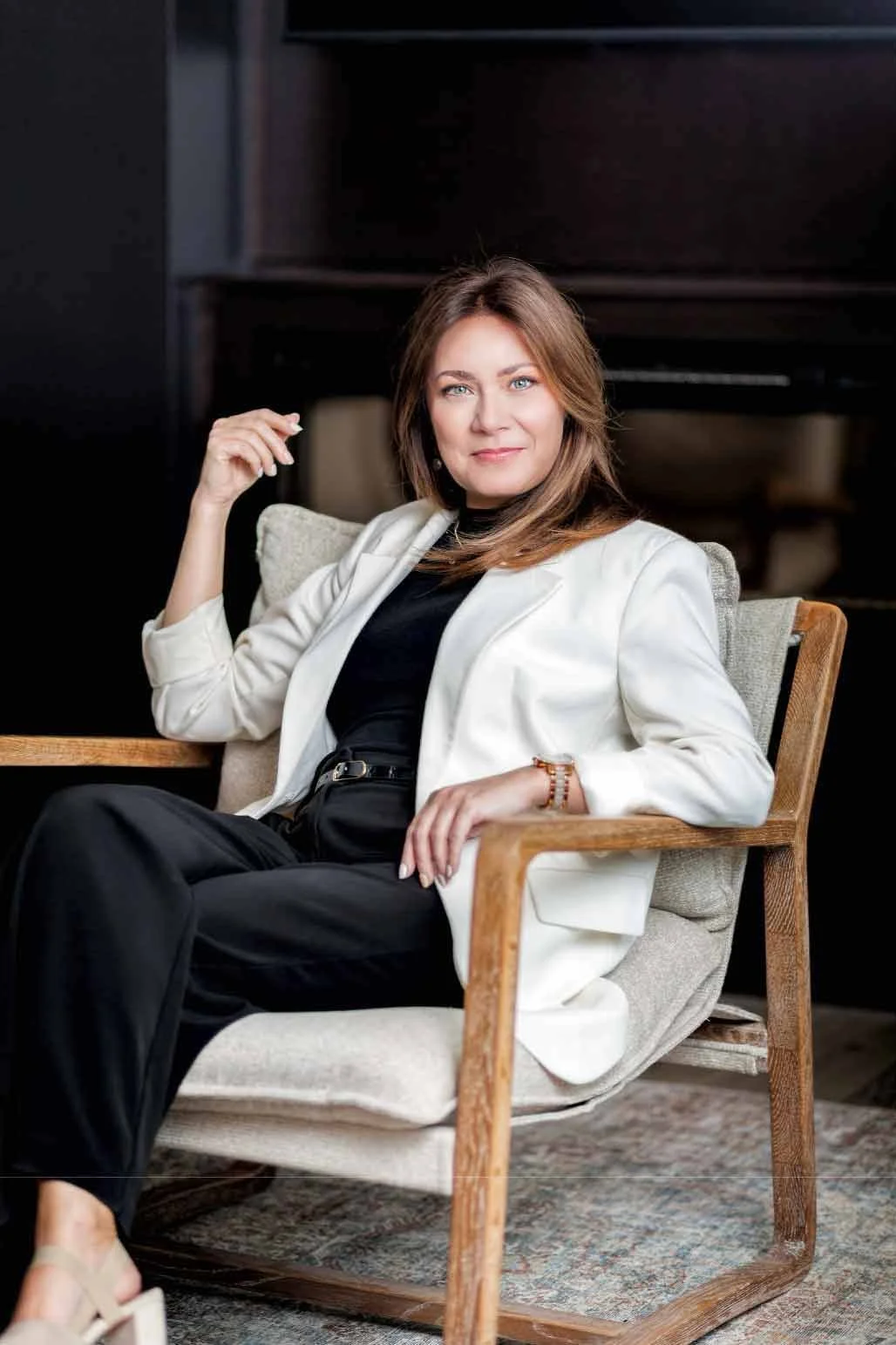 A woman with brown hair and blue eyes wearing a white blazer, black top, and black pants, sitting on a beige armchair with wooden arms in a room with dark background.