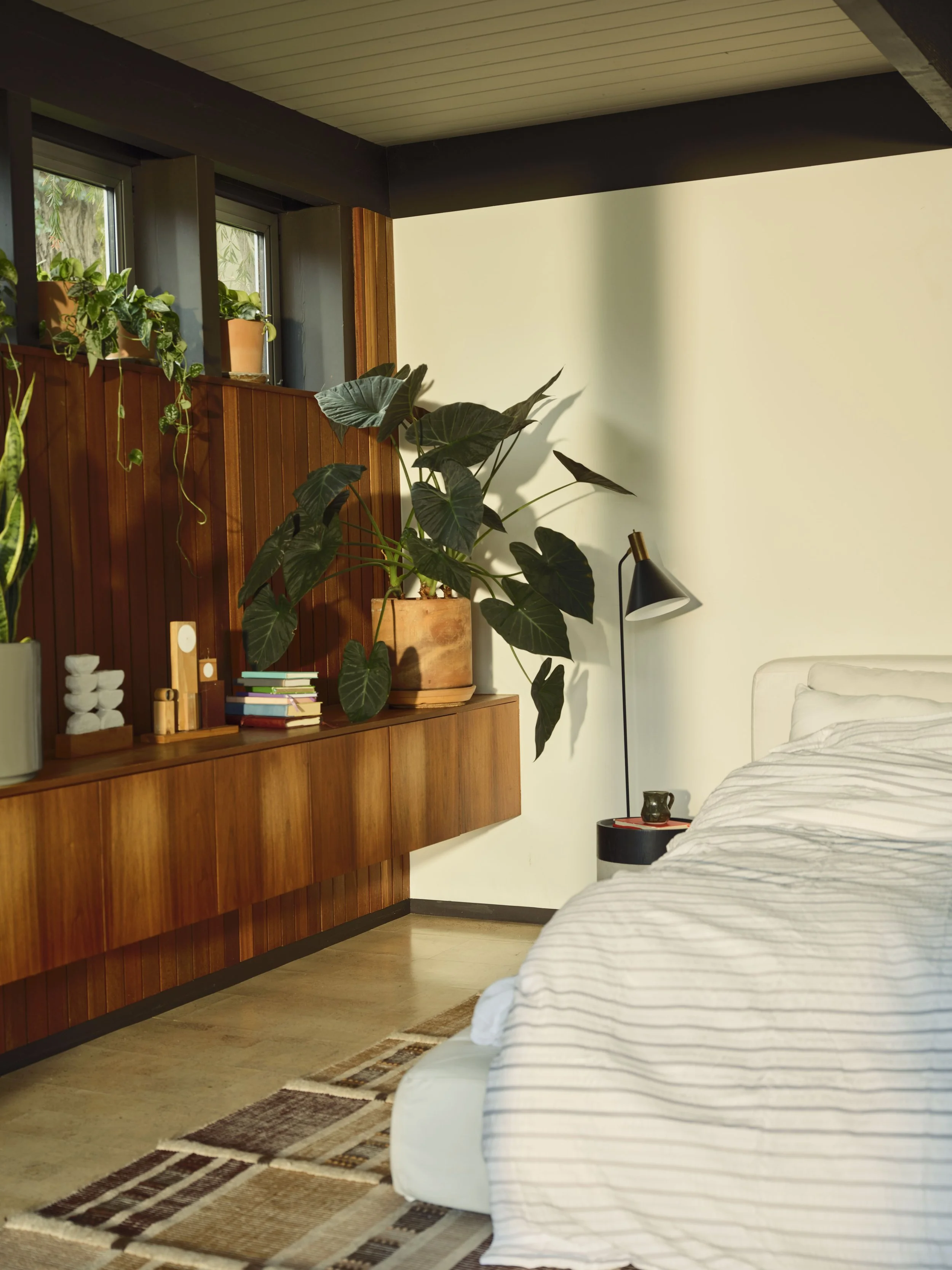 A cozy bedroom with a bed, some potted plants, a wooden sideboard, and a black floor lamp, with a cream-colored wall and a small window with additional plants.
