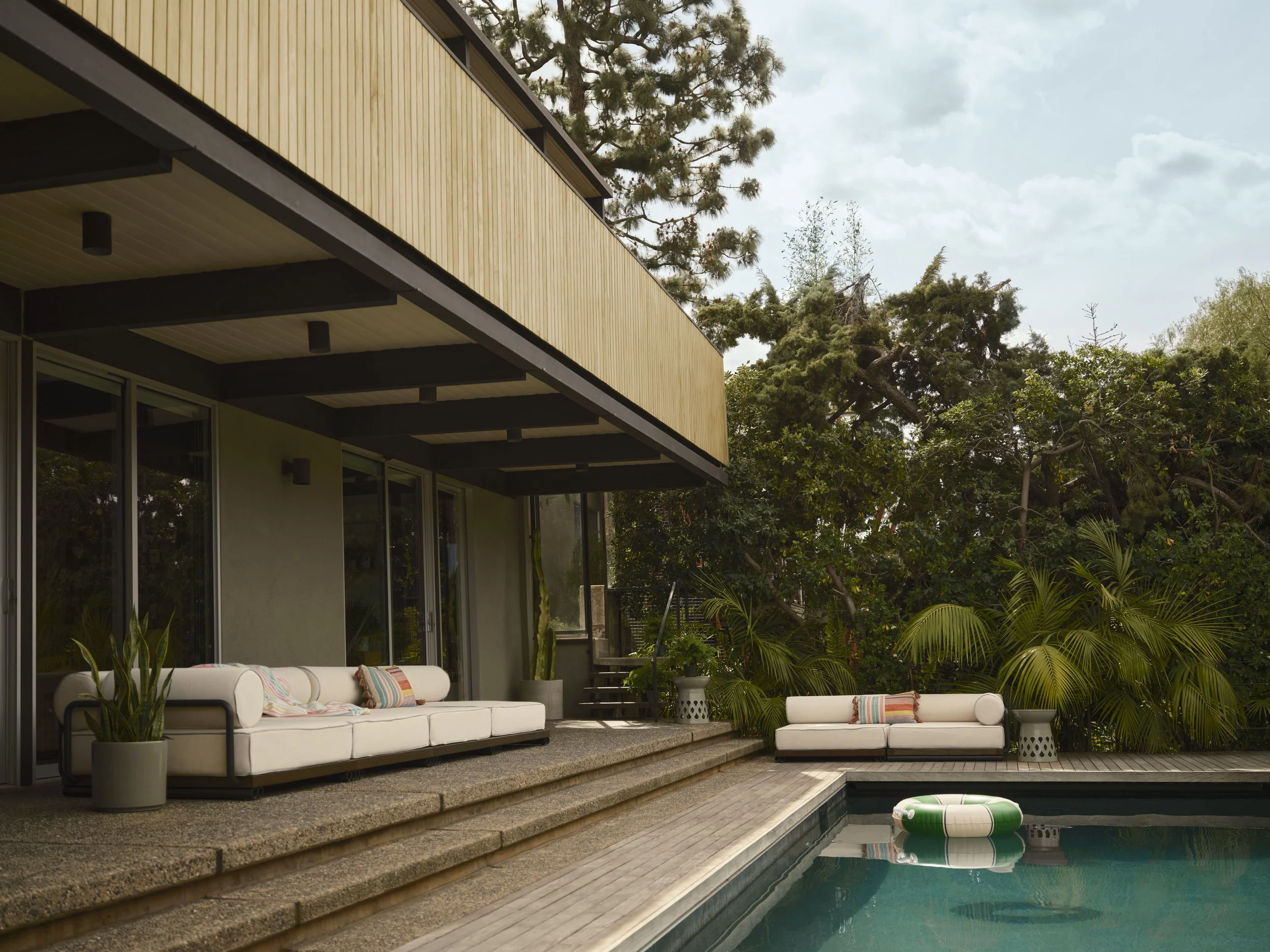 An outdoor patio area with white sofa and chair, potted plants, and a swimming pool with inflatable float, surrounded by trees and a modern building with large glass windows.