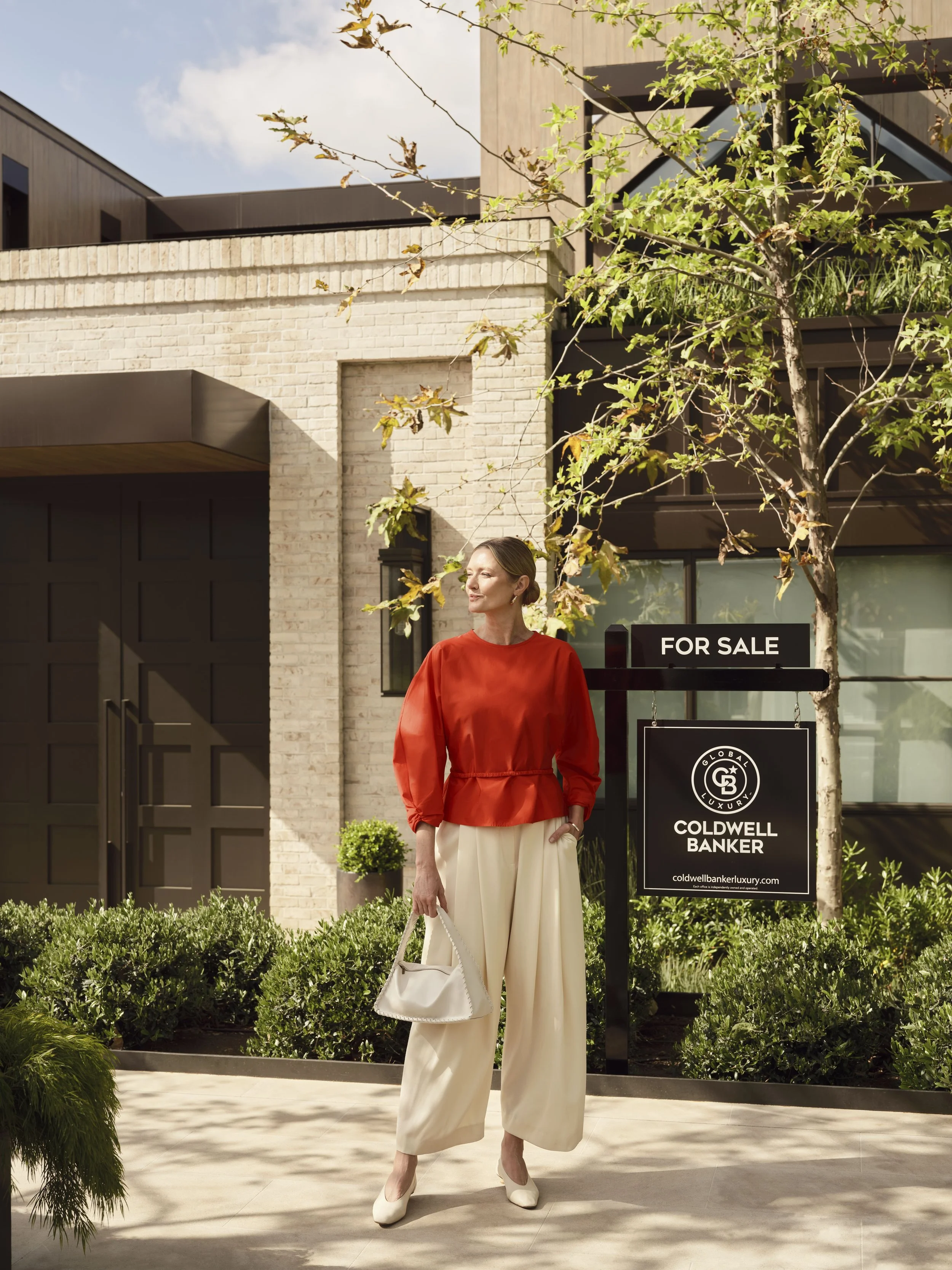 A woman in a red blouse and cream-colored wide-leg pants standing outside near an Coldwell Banker Sign