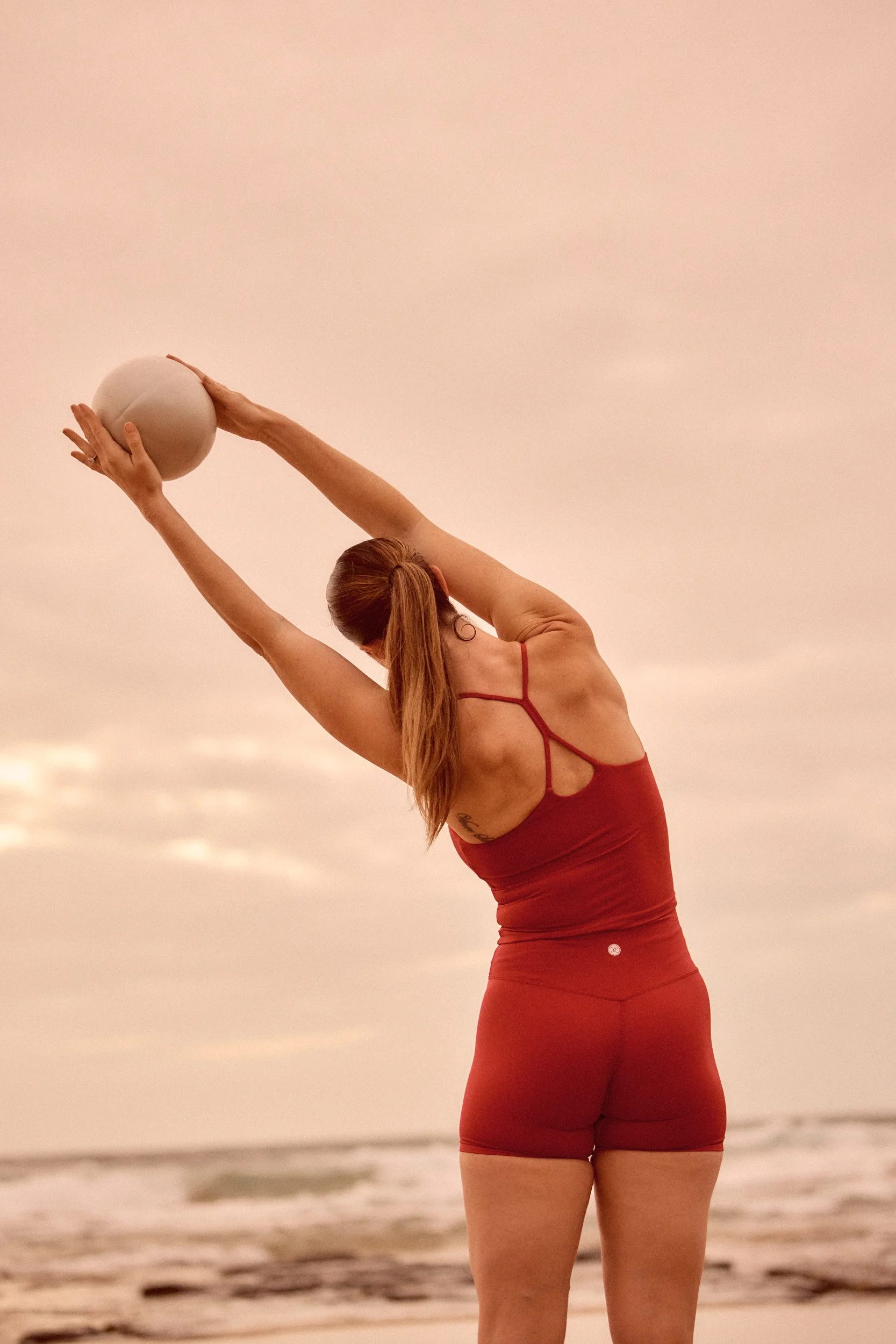 Jade from Motion Pilates in a red sports bra and matching shorts stretching with a pilates ball at Austinmer beach doing a pilates session during sunset, holding a beige ball above her head.