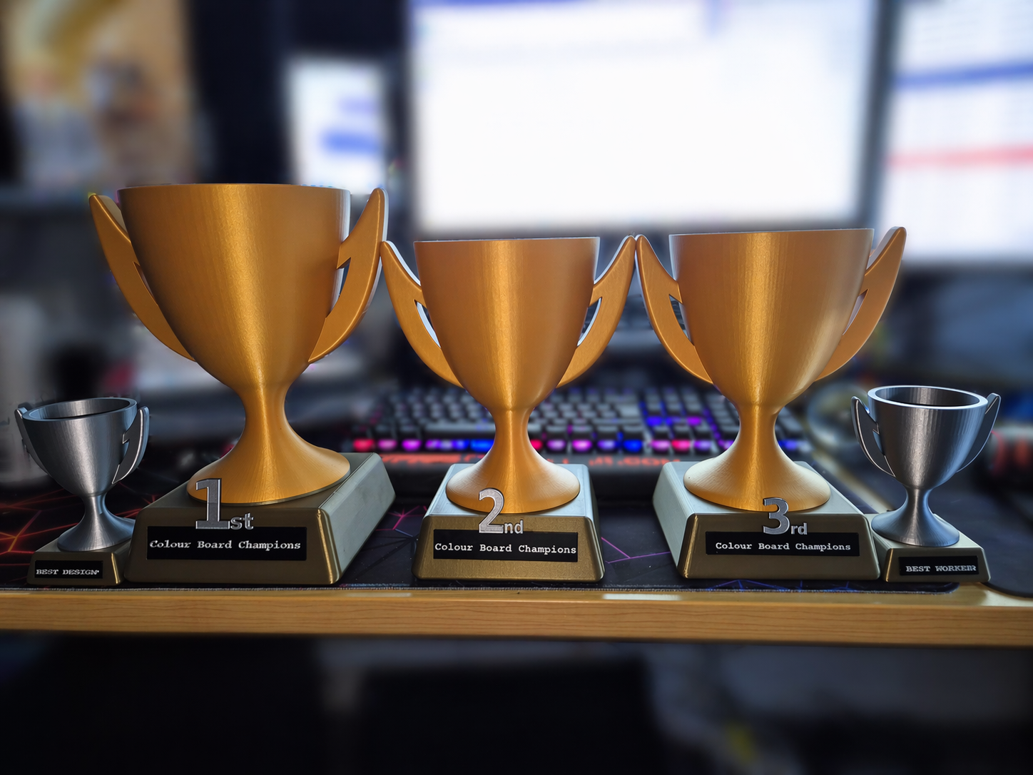 Four trophies on a table, with three large gold trophies labeled 1st, 2nd, and 3rd place, and two smaller silver trophies labeled Best Design and Best Worker.