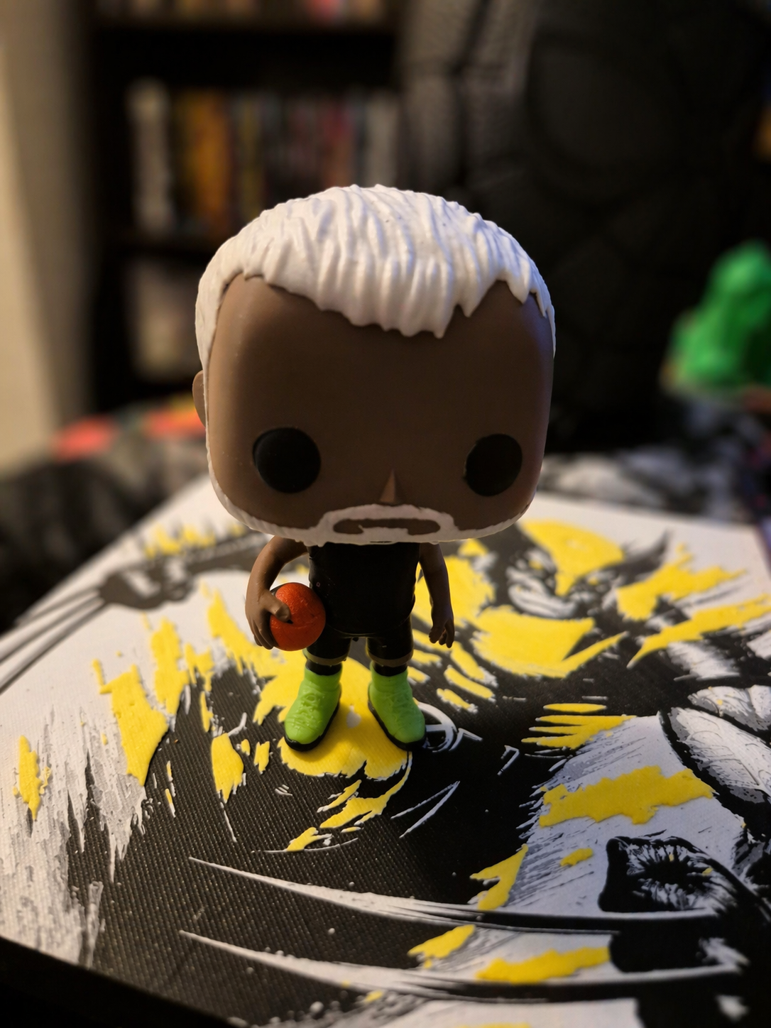 A Funko Pop! figurine of a male baseball player with white hair, brown skin, and a beard, holding a red baseball, wearing a black uniform with green shoes, standing on a black and yellow art piece.