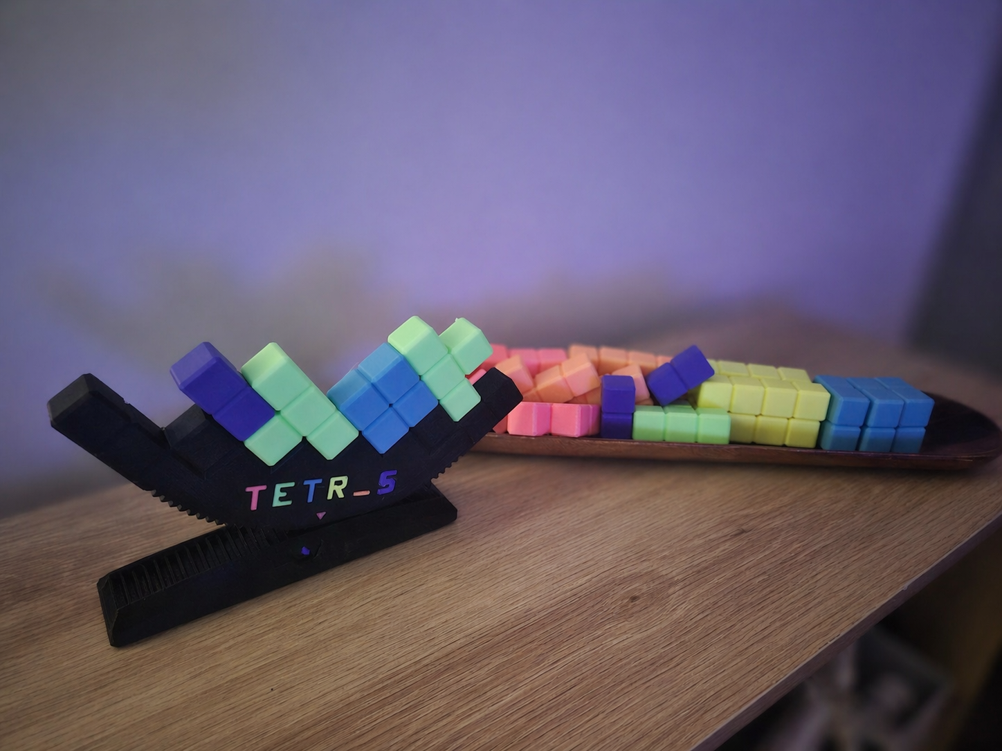 Colorful 3D-printed Tetris blocks in black stand and on a wooden tray, on a wooden table with a purple background.
