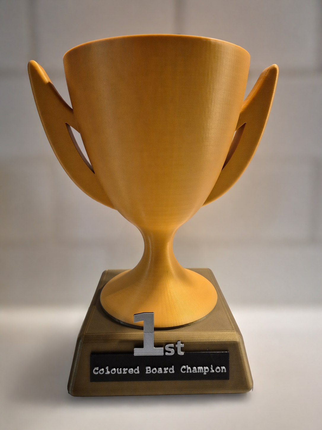 A gold-colored trophy with a cup shape, mounted on a base with a plaque reading "1st Coloured Board Champion."