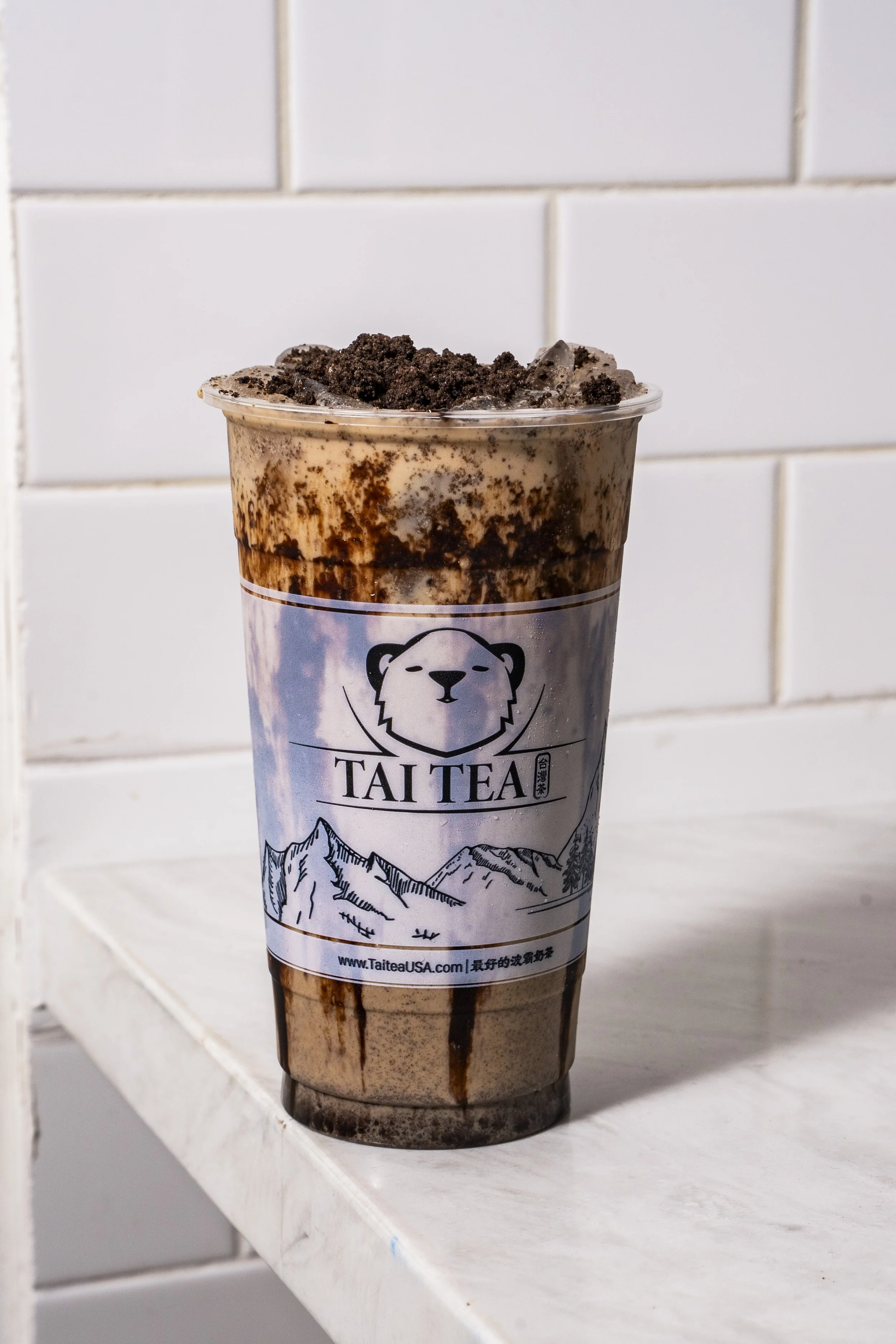 A tall plastic cup of chocolate milk tea topped with crushed cookies or Oreos, with chocolate syrup drizzled inside the cup and on the surface of the drink, placed on a white shelf against a white tiled wall.