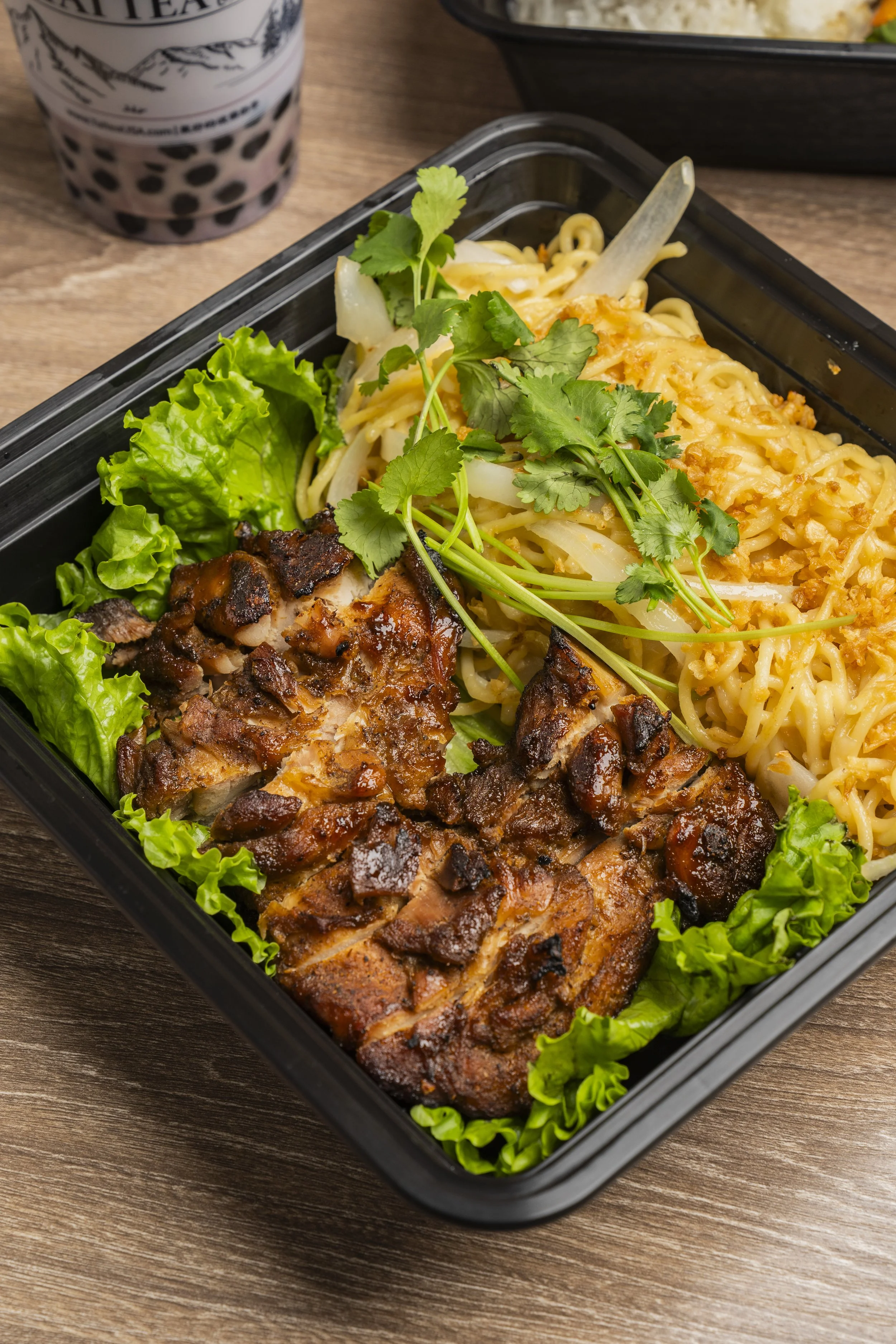 A black container with grilled chicken, lettuce, noodles, and cilantro, placed on a wooden table with a drink in the background.