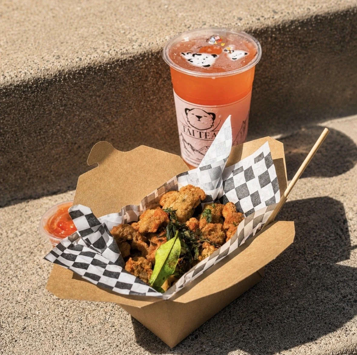 Fried chicken pieces with green herbs in a cardboard tray lined with checkered paper, a small cup of ketchup, and a pink drink with panda and bear images, on a concrete surface.