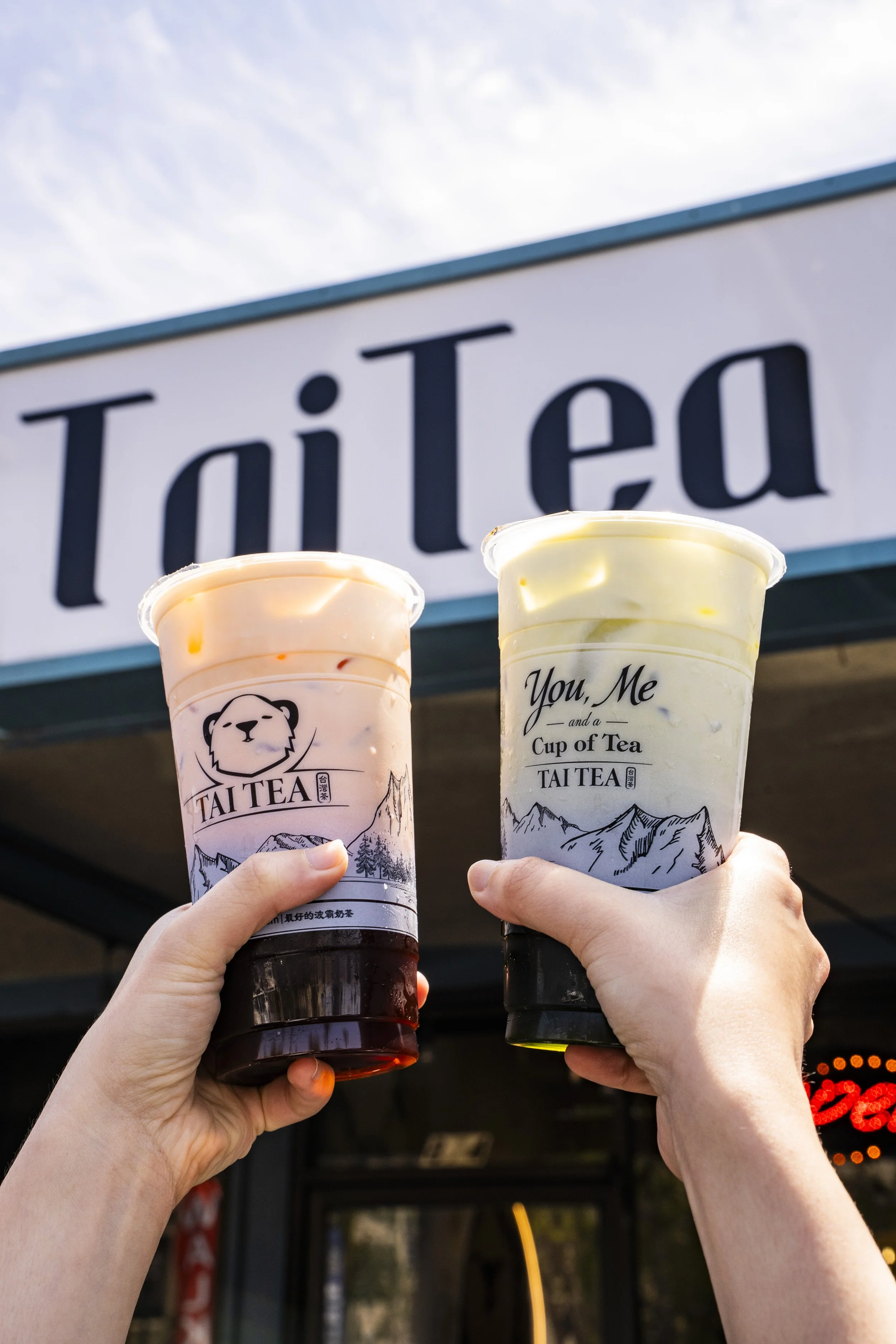 Two hands holding cups of bubble tea in front of a Tai Tea sign. One cup has pink-colored tea, and the other has yellow-colored tea.