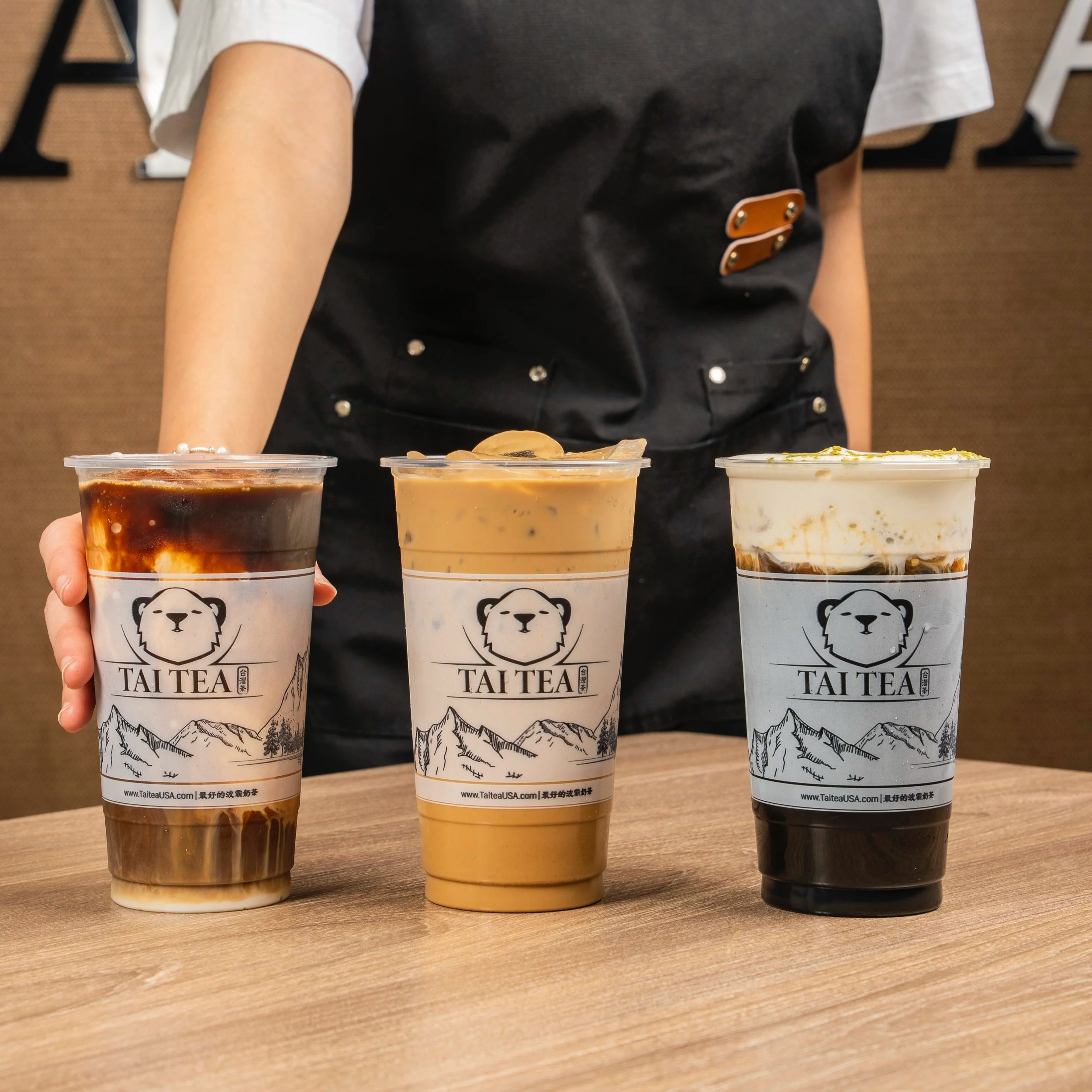 Three large cups of bubble tea with different flavors on a wooden table, featuring a person in a black apron in the background.