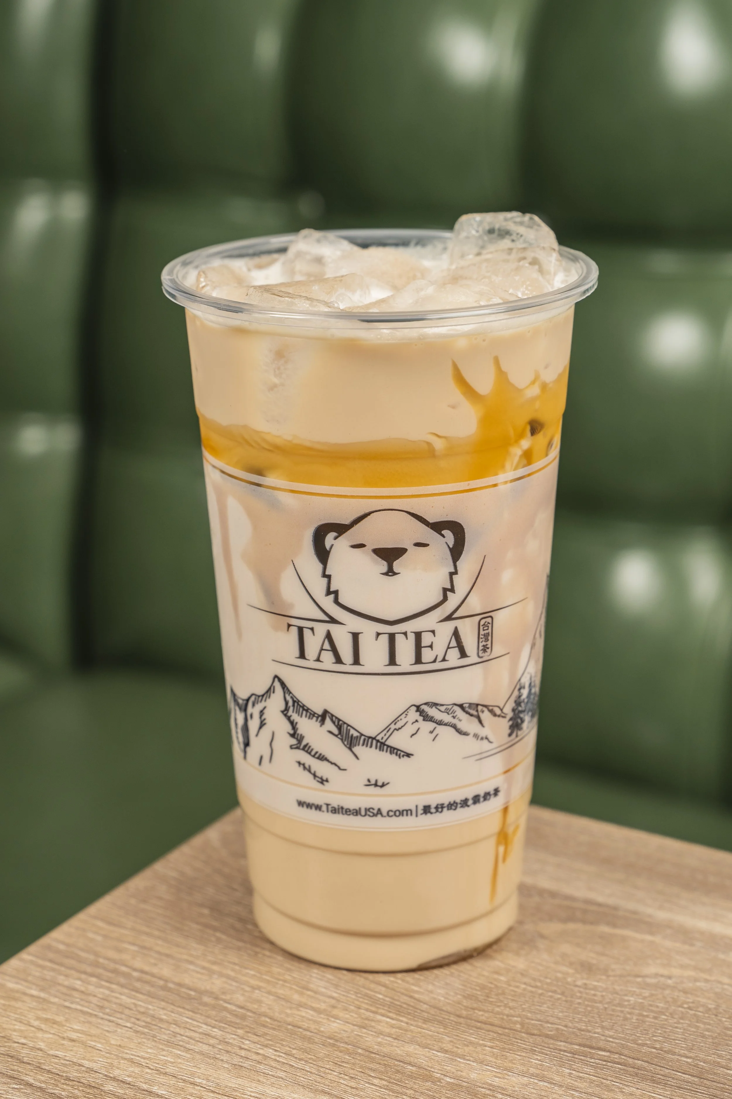 A plastic cup filled with iced tea, with layers of cream and caramel, on a wooden table. The cup has a 'Tai Tea' logo featuring a bear's face and mountain illustrations.