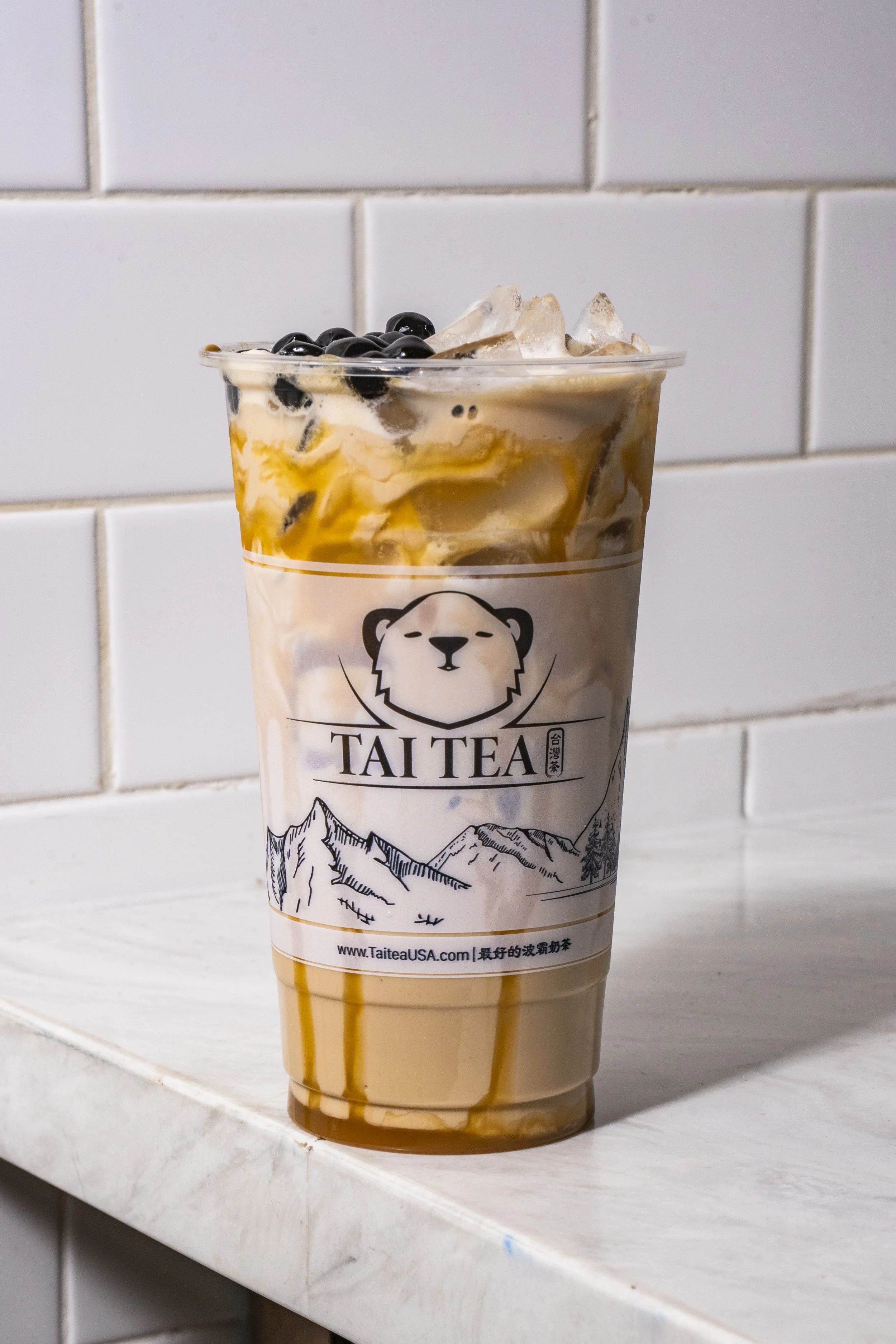 A large to-go cup from Tai Tea with tapioca pearls on top and caramel syrup inside. The drink appears to be a caramel milk tea with ice, placed on a white marble countertop against a white tiled wall.