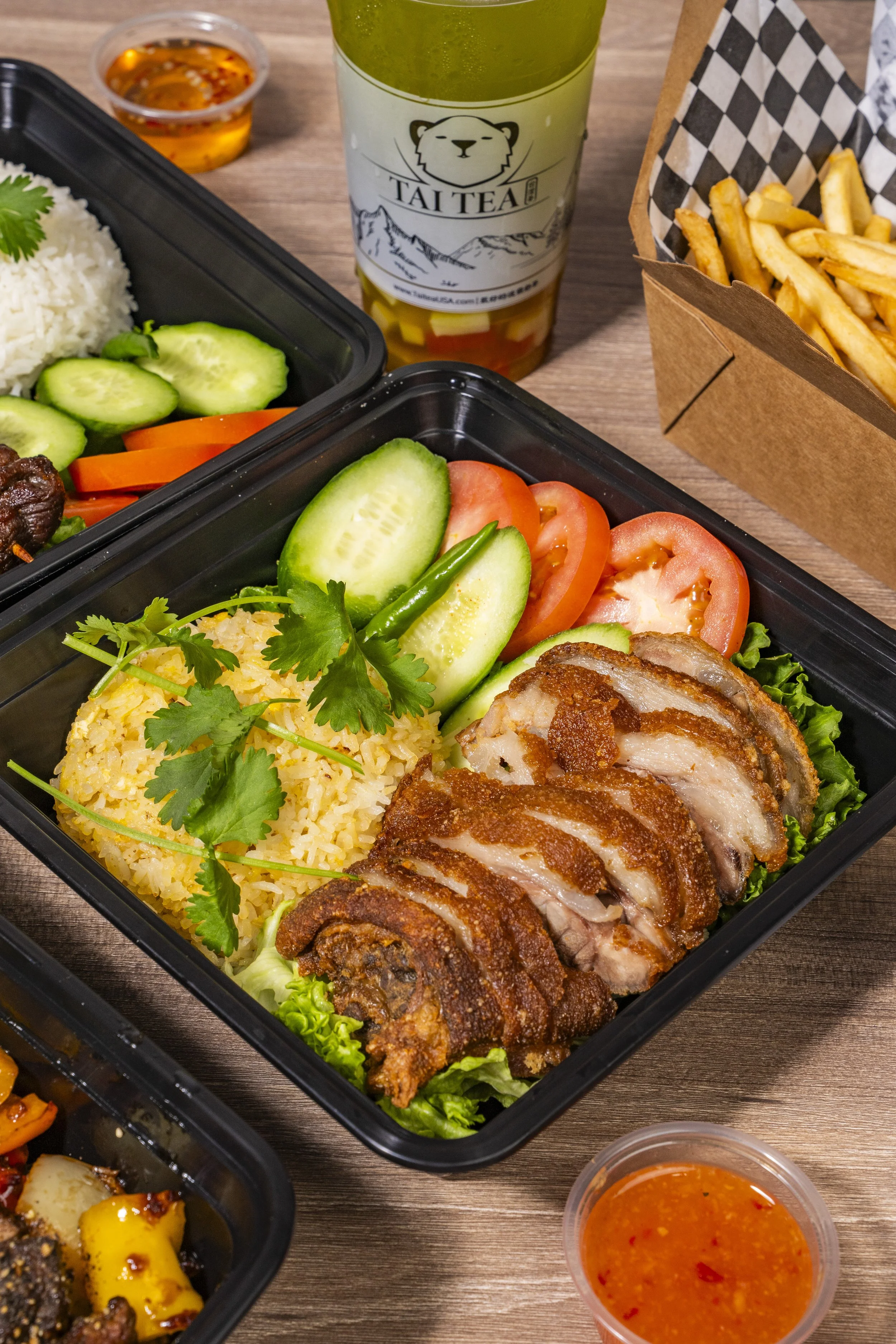 A bento box filled with sliced fried chicken, cucumber, tomato, rice, and cilantro, with a side of French fries, a cup of spicy dipping sauce, and a bubble tea in a plastic cup with a straw.