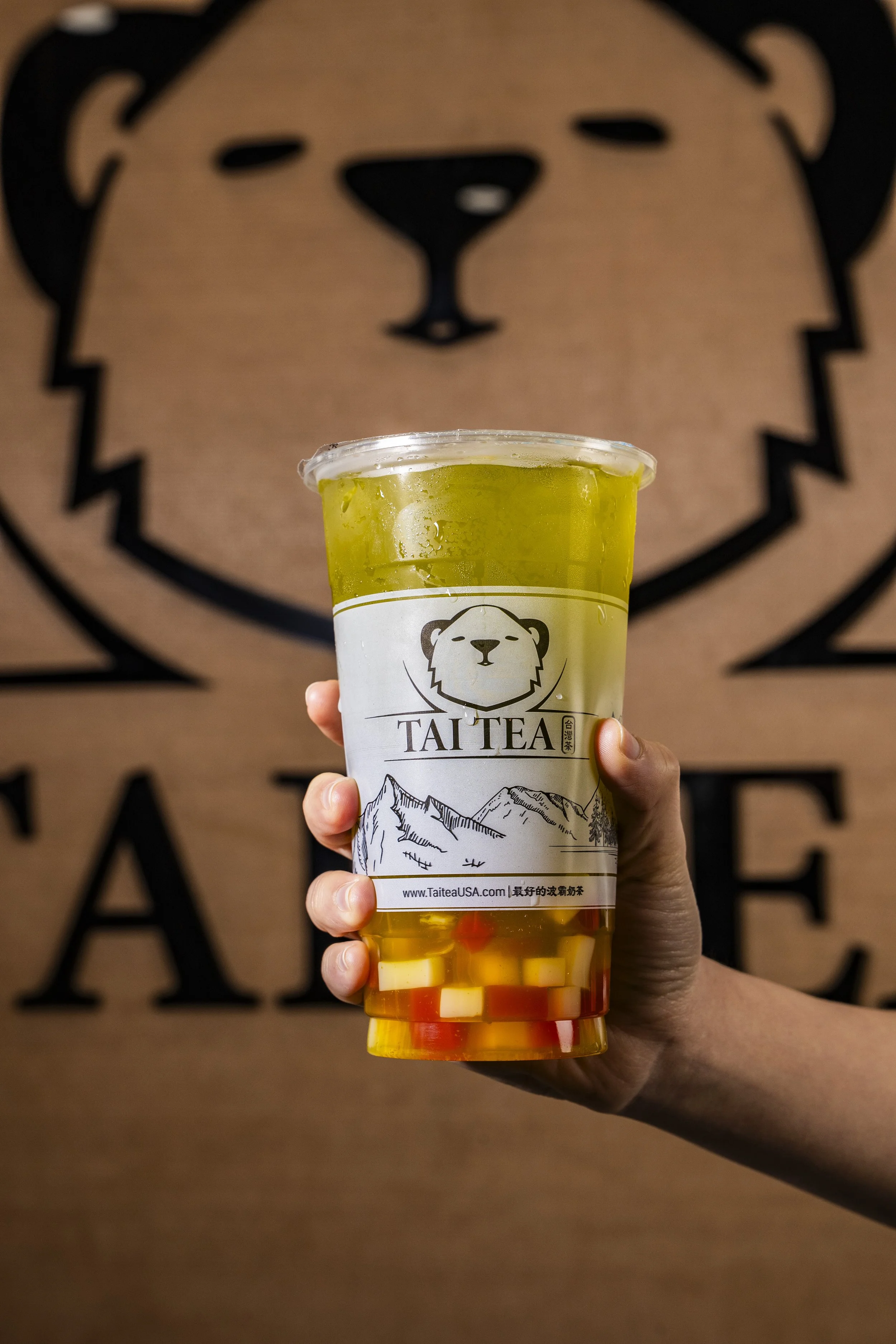 A hand holding a cup of bubble tea with yellowish liquid and colorful cubes at the bottom, with a branded label featuring a bear's face and mountain scenery, in front of a large bear logo on a wall.