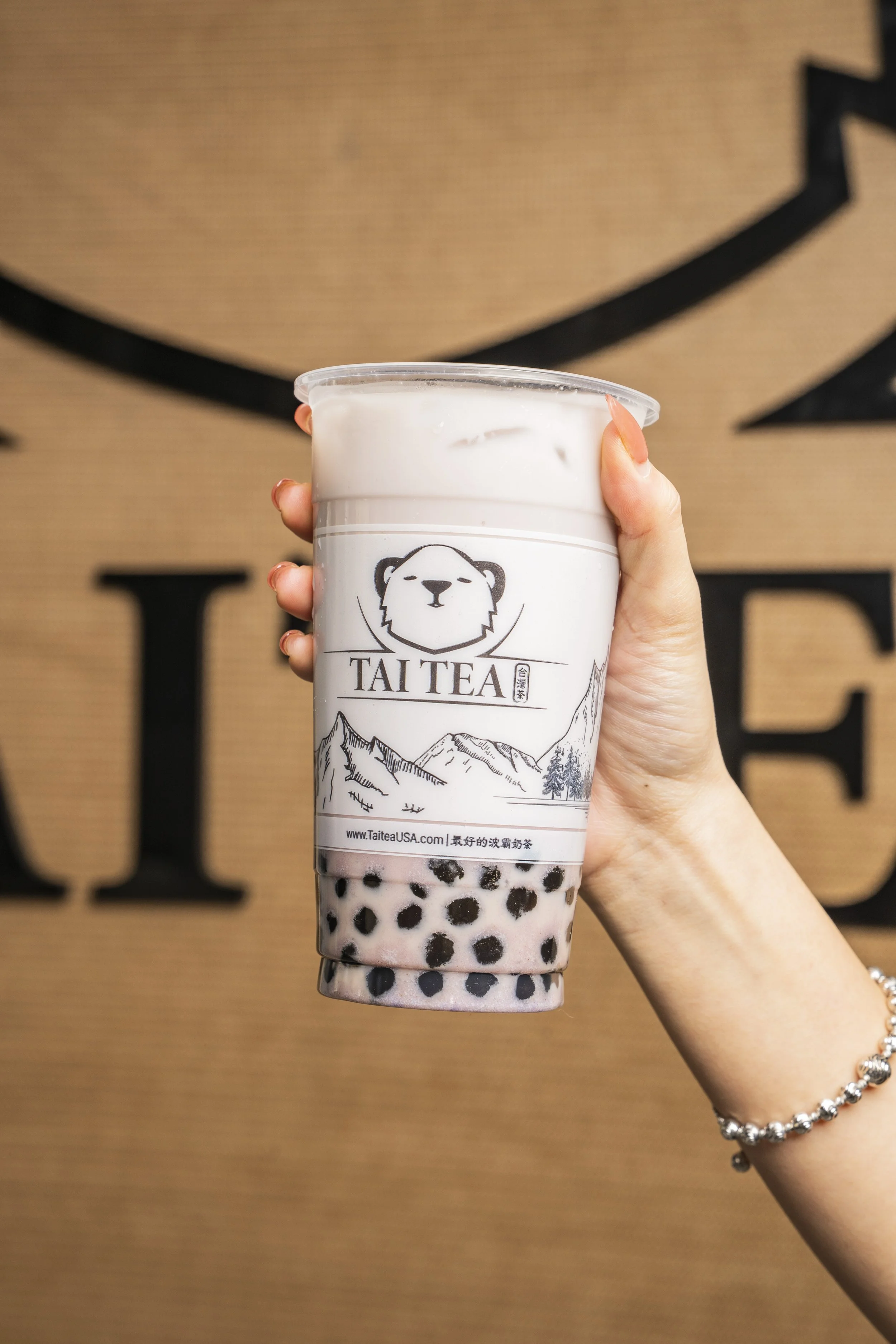 A hand holding a cup of bubble tea from Tai Tea, with black tapioca pearls at the bottom, in front of a decorative background with large black letters.