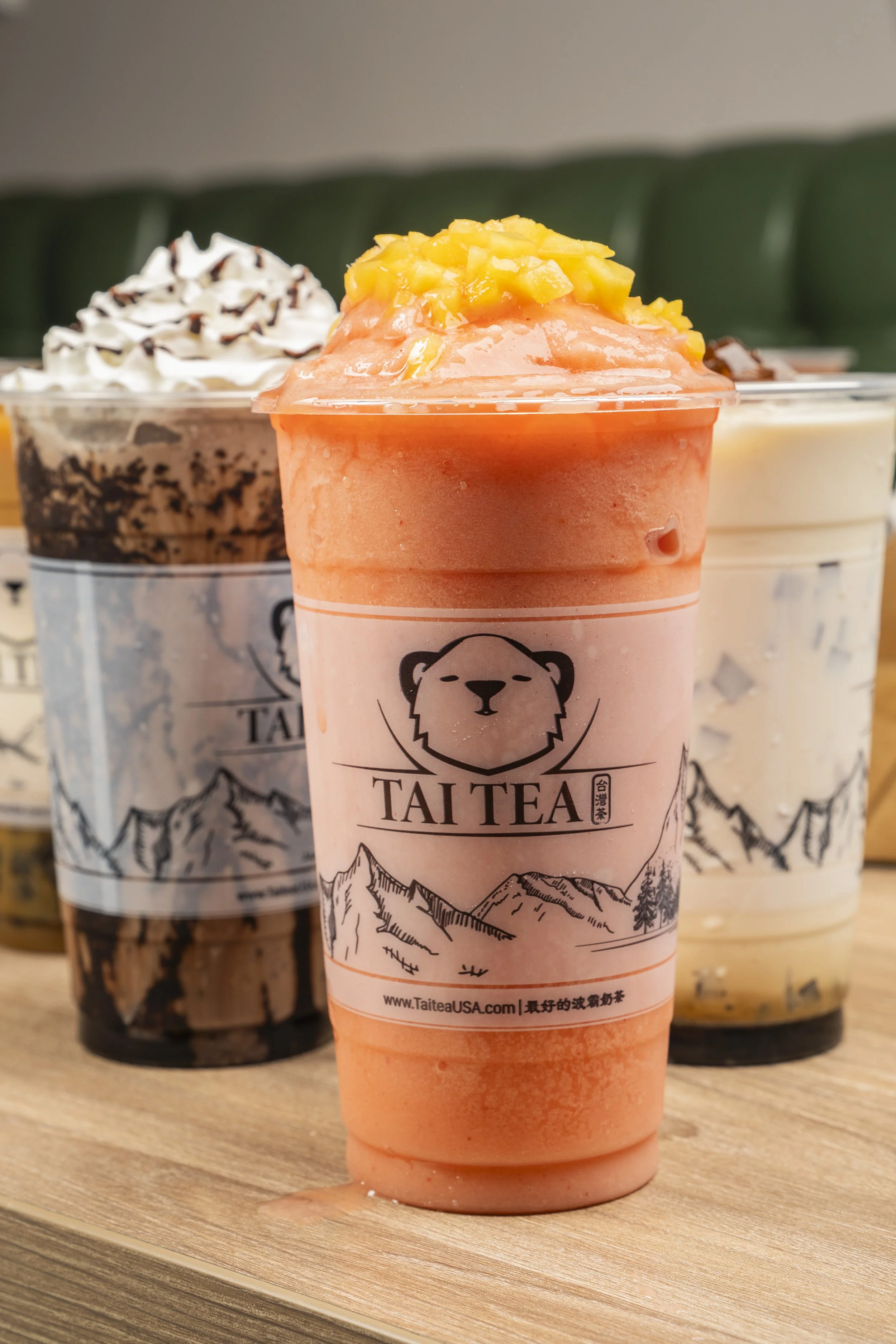 A pink mango-flavored blended drink with chopped mango on top, from Tai Tea, on a wooden surface, with bluered drinks containing whipped cream and chocolate toppings in the background.