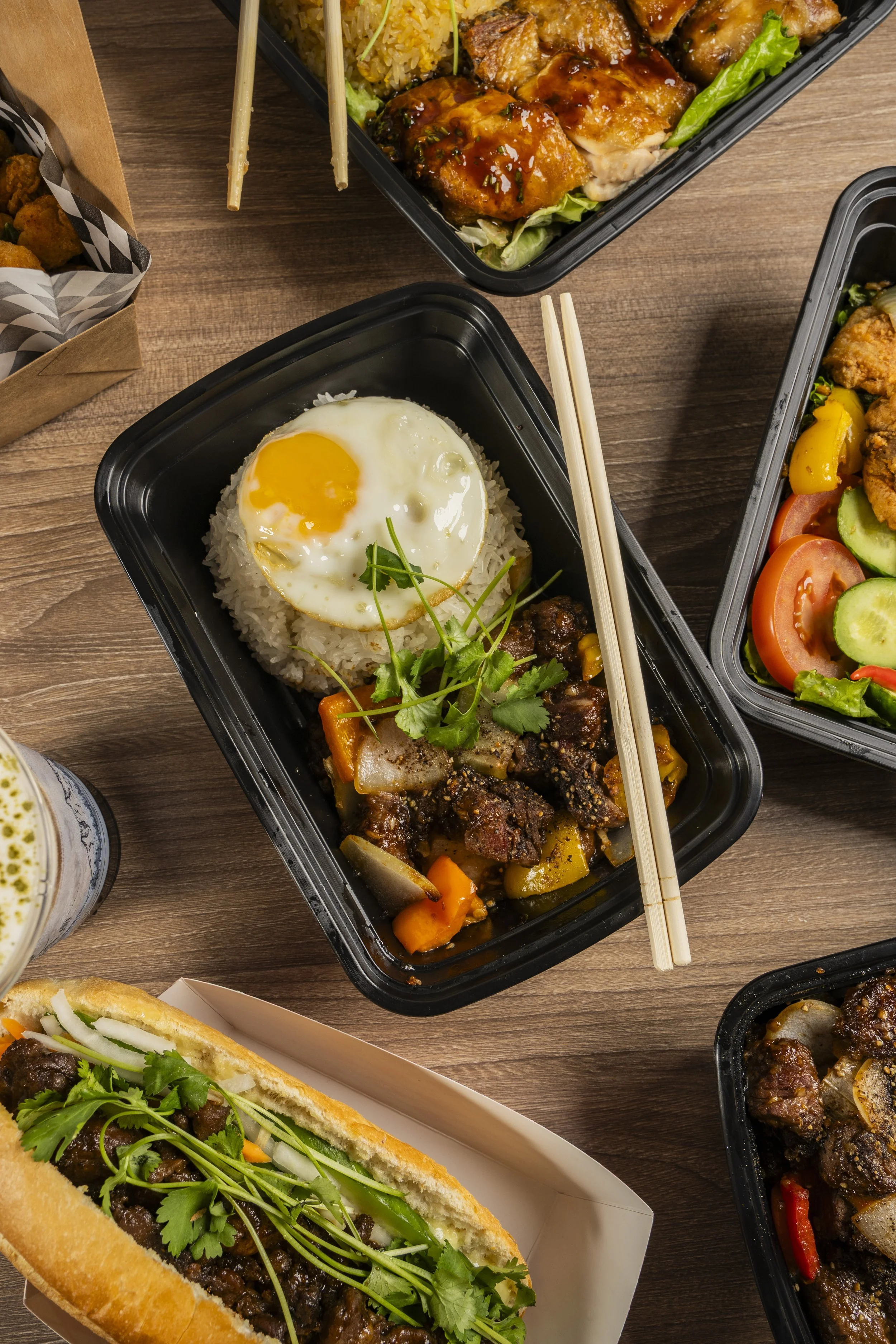 Various Asian-inspired dishes in black takeout containers on a wooden table, including rice with a fried egg and beef stew, and other dishes with vegetables and meat.