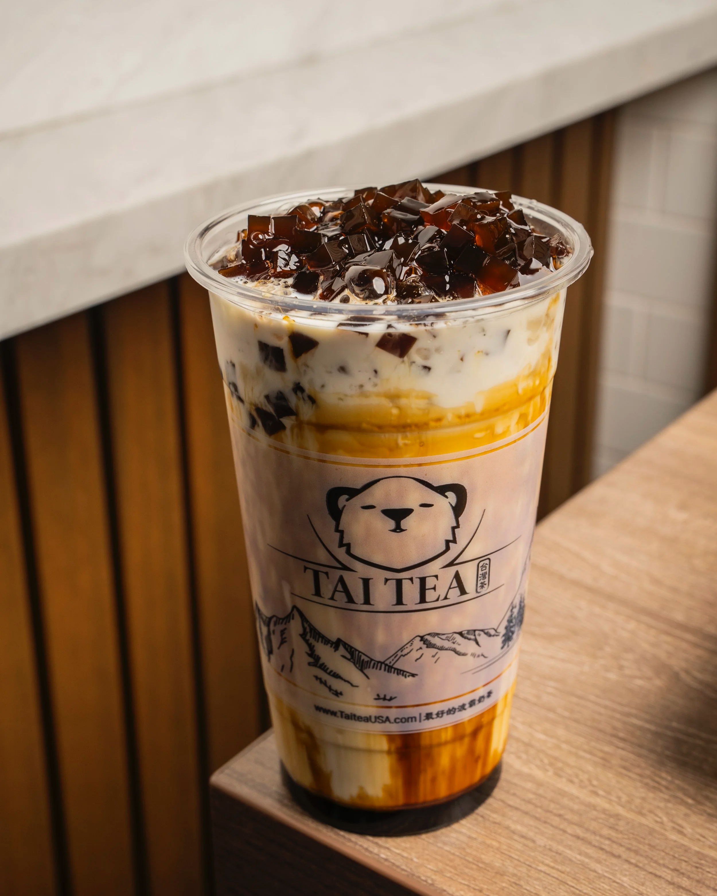 A large cup of bubble tea with black tapioca pearls and caramel syrup, topped with ice and cream, from Tai Tea, on a wooden table.