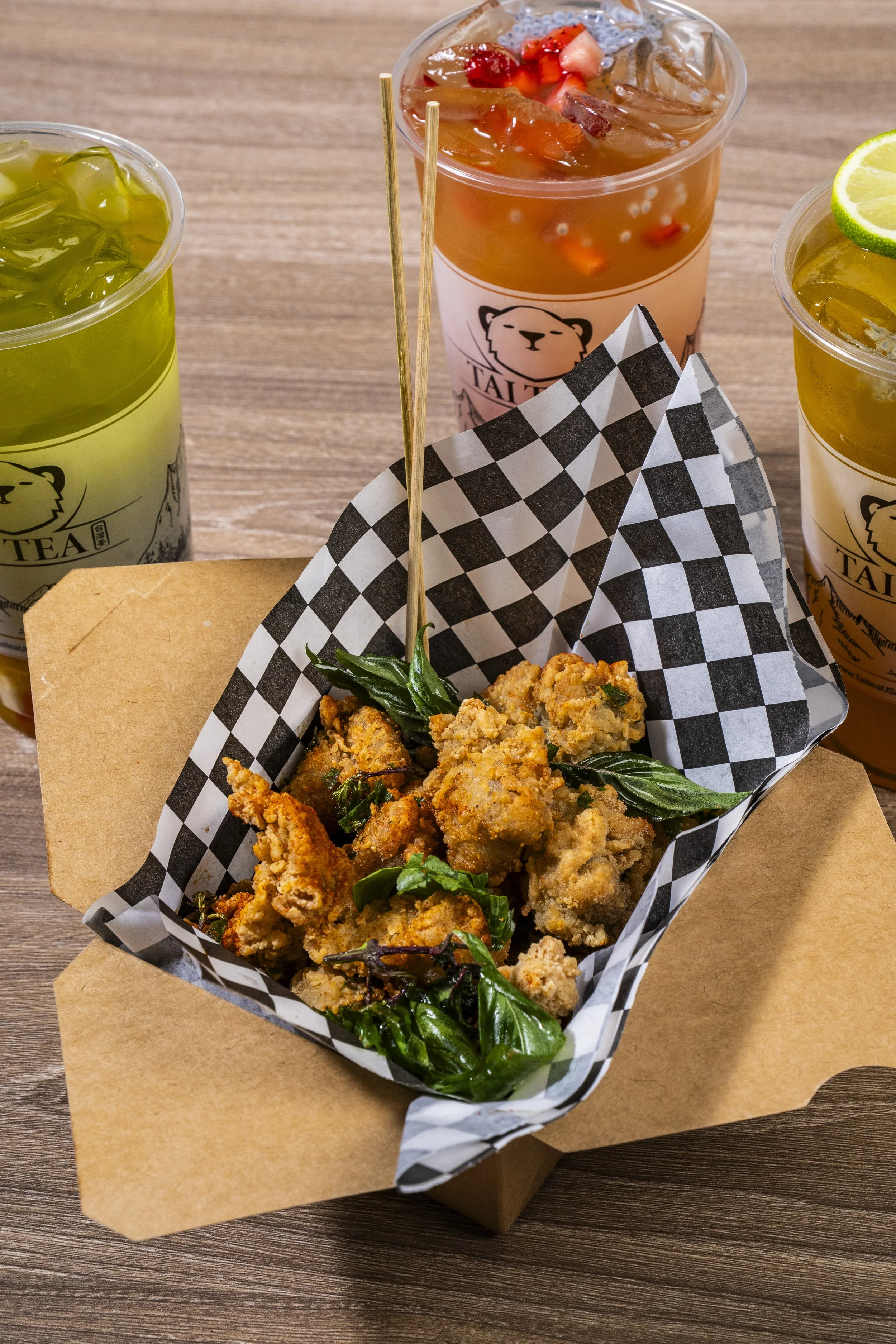 Fried chicken pieces garnished with basil leaves in checkered paper basket, with three iced beverages in plastic cups with lime and berry garnishes on a wooden table.