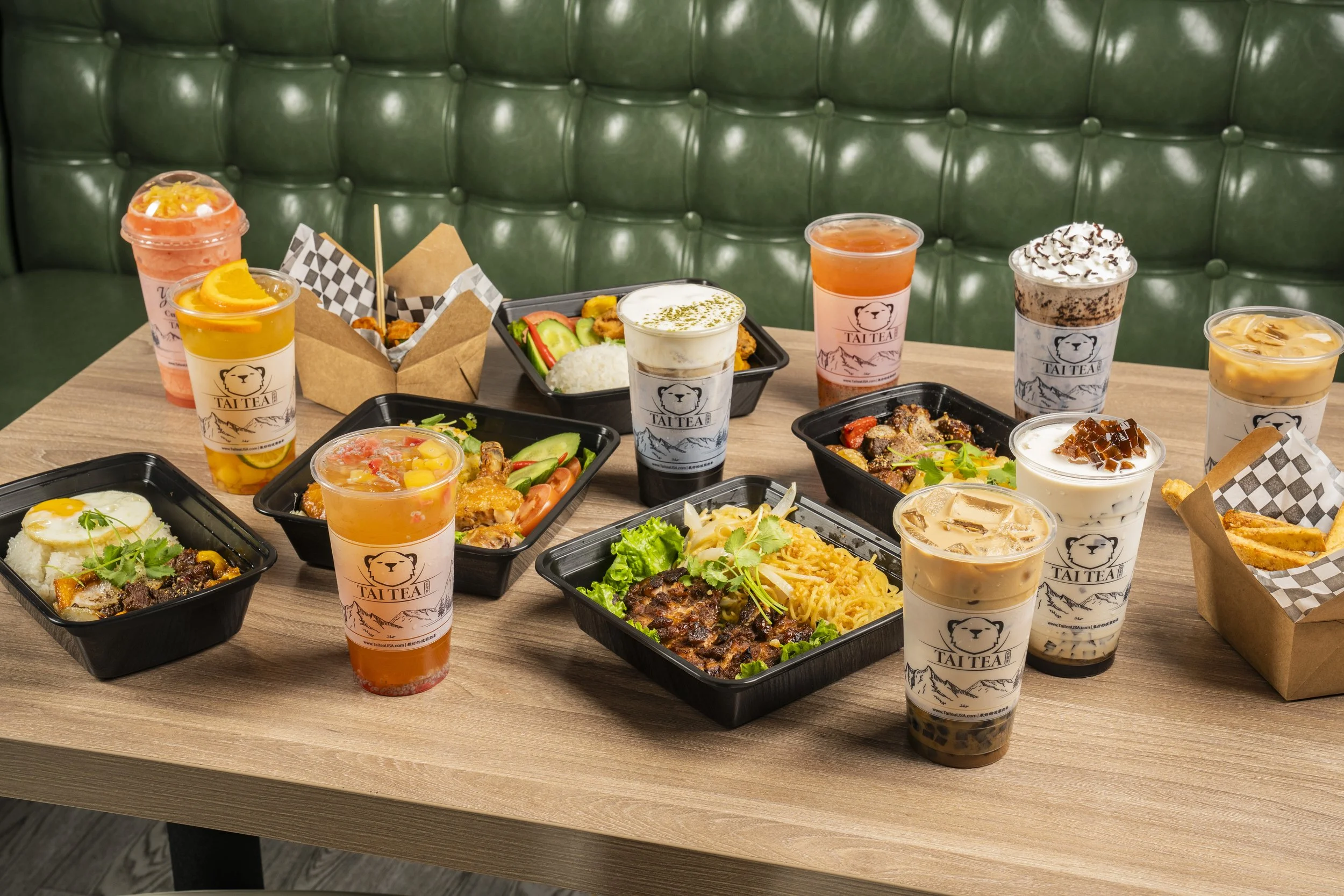 A table filled with various Asian-style takeaway food containers and drinks, including bubble tea and smoothies, on a wooden table with a green upholstered booth in the background.