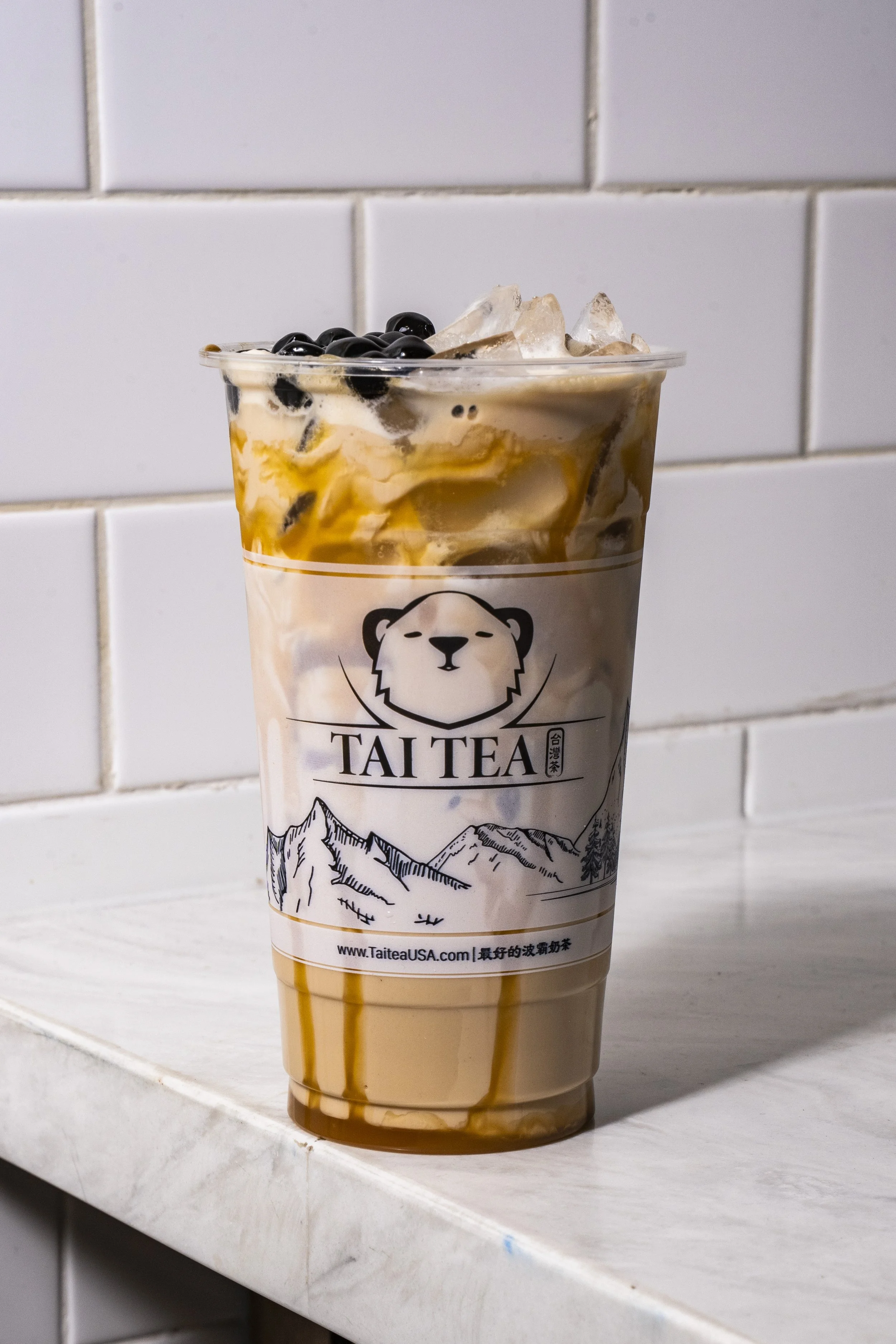 A large plastic cup of bubble tea with black tapioca pearls, ice, cream, caramel drizzle, and a mountain graphic on the sleeve, placed on a white tiled shelf.