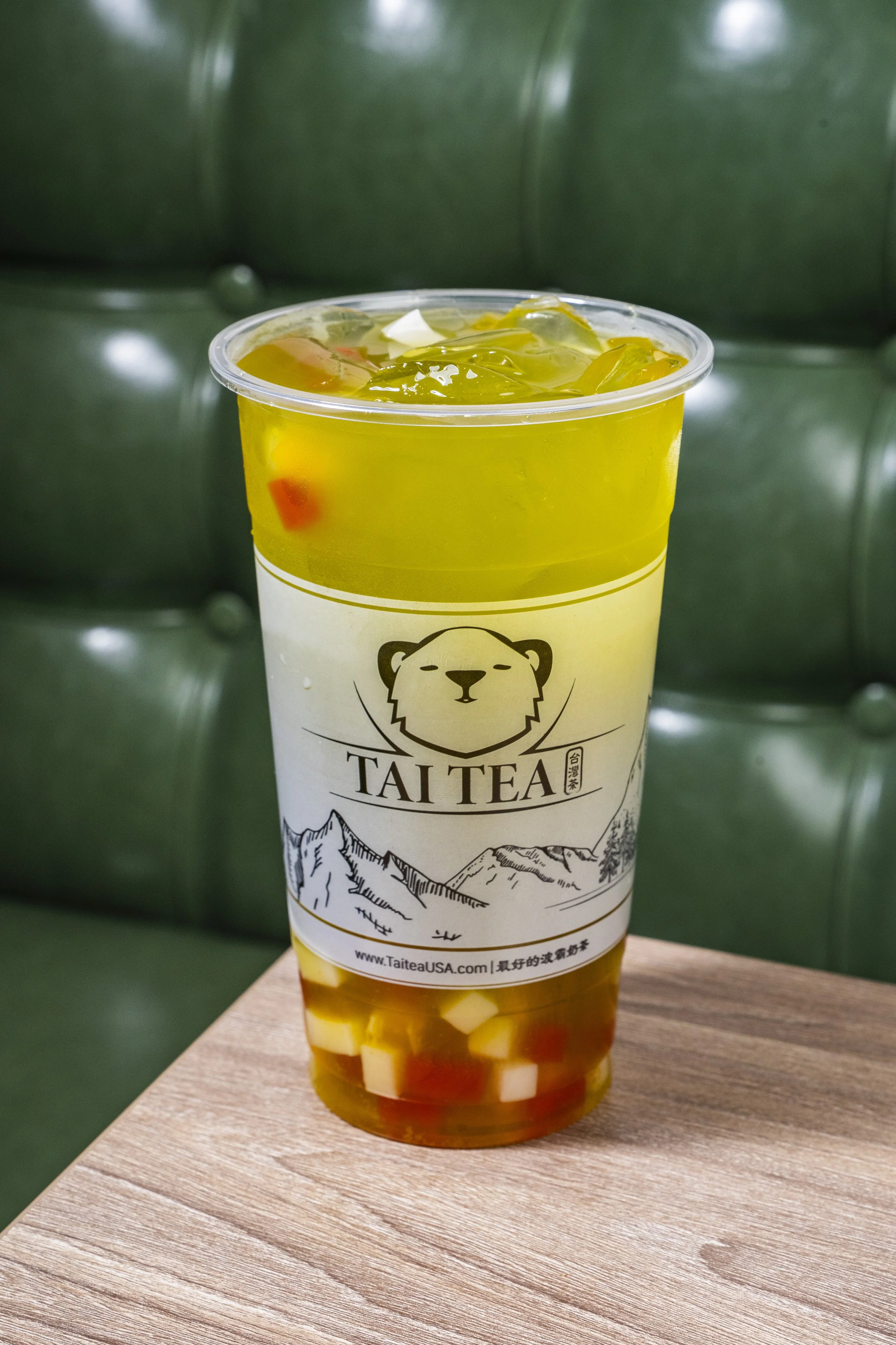 A cold beverage in a clear plastic cup with a mountain illustration and a logo featuring a bear, placed on a wooden surface with a dark green cushioned background.