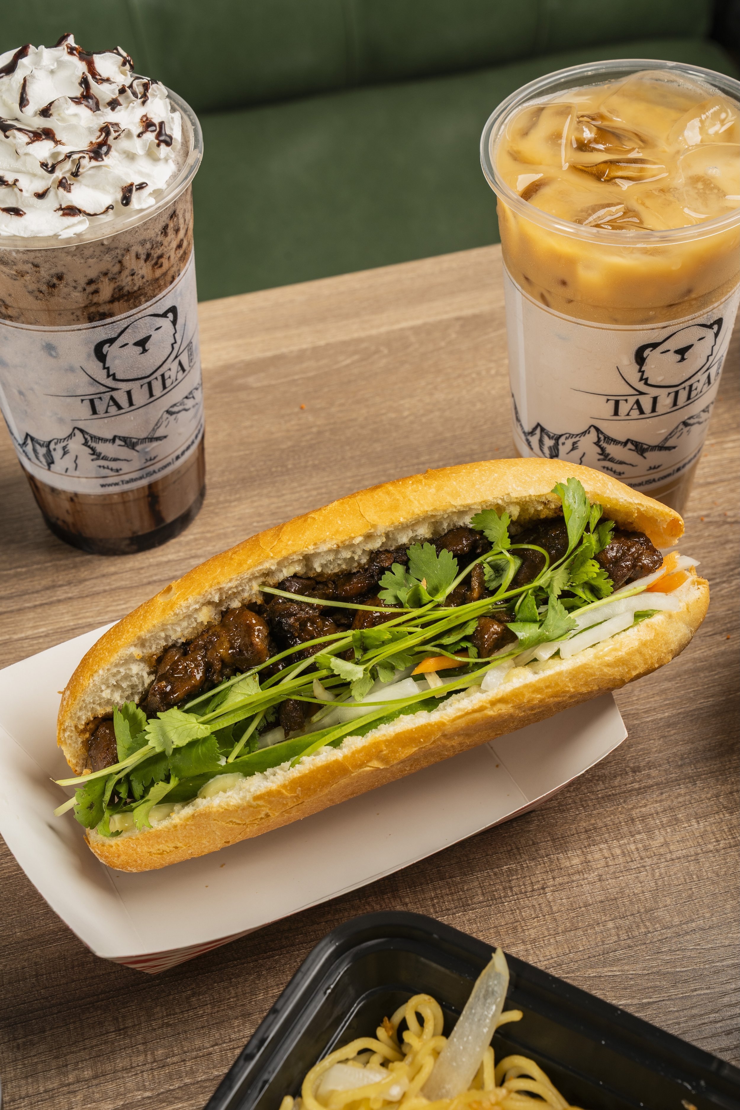 A takeout meal featuring a hoagie sandwich with steak, lettuce, and cilantro, two iced drinks from Tai Tea with whipped cream and chocolate syrup and iced coffee with creamer, and a container of noodles with vegetables.