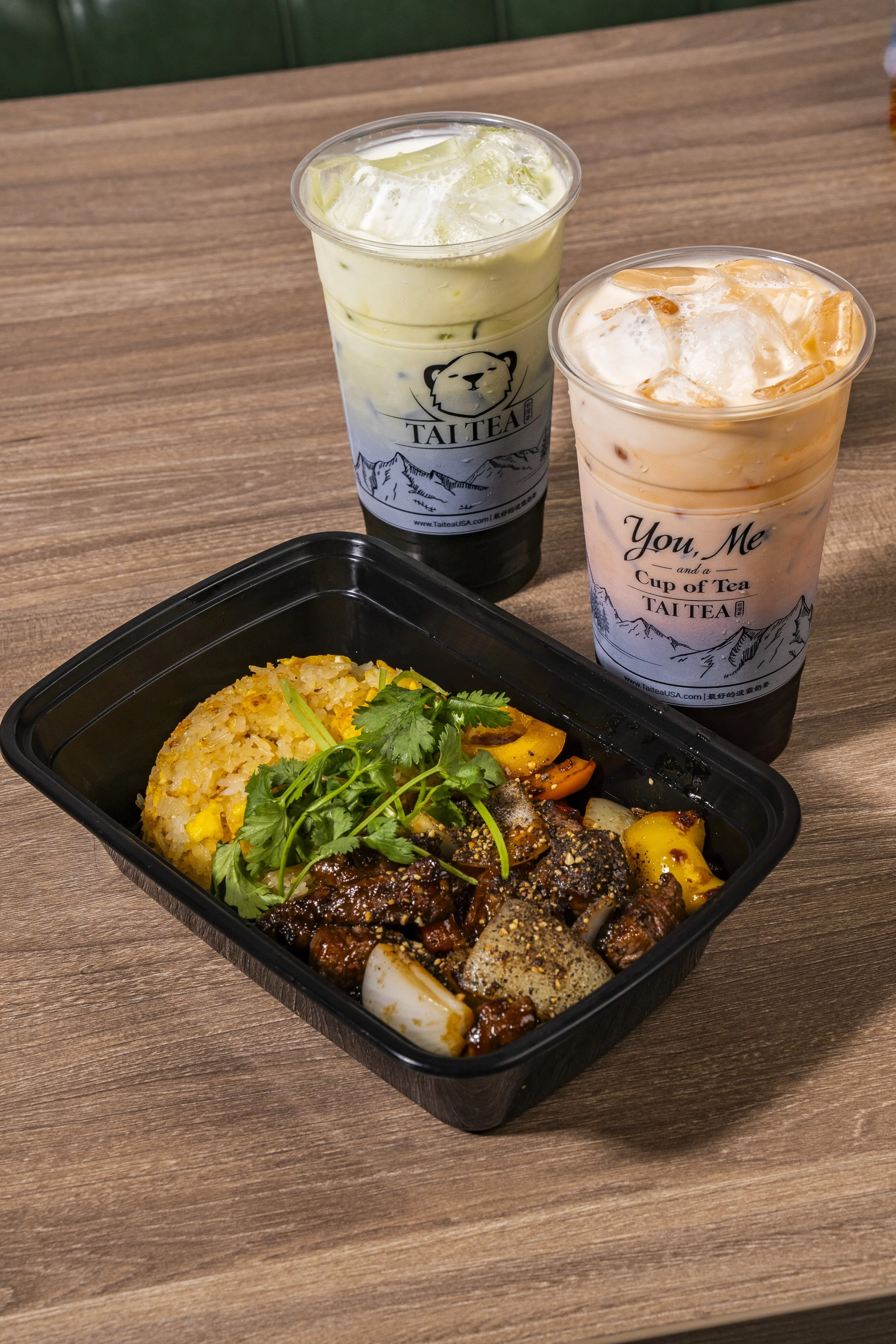 Two cups of iced tea, one with a greenish hue and the other with an orange hue, on a wooden table, with a black takeout container of fried rice and beef stir-fry garnished with cilantro in the foreground.