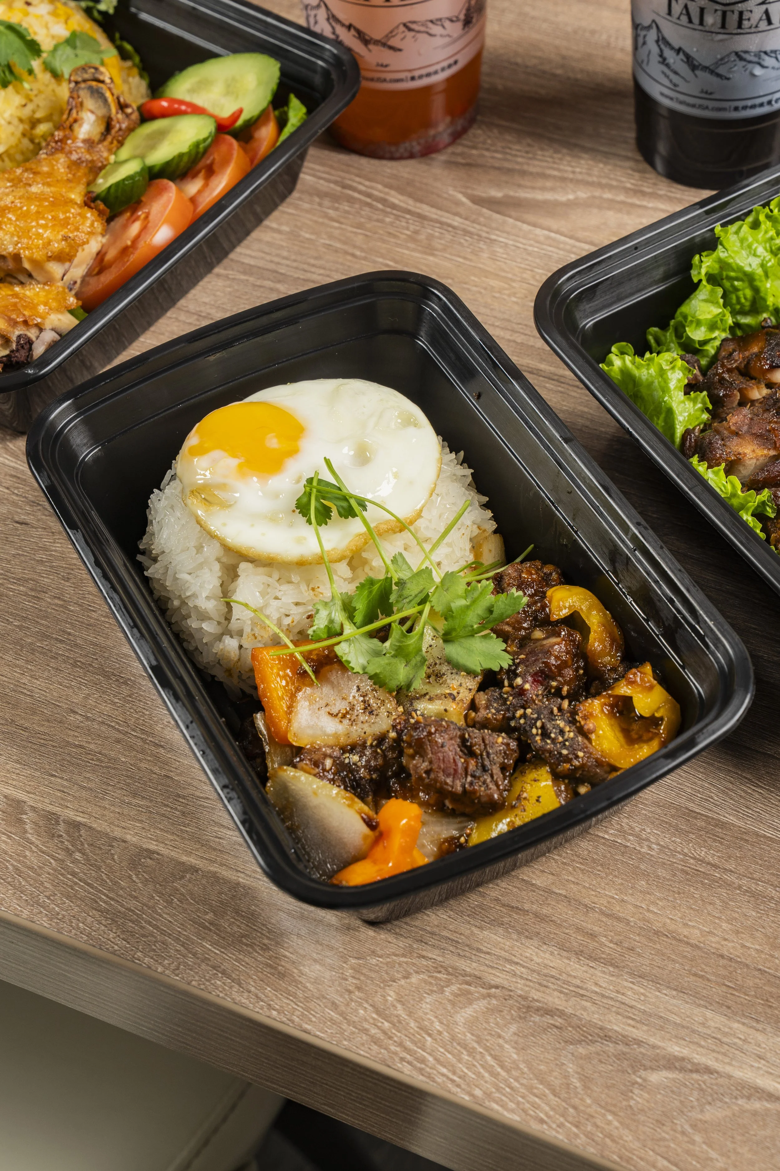 Black plastic meal container with rice, a fried egg, beef stew with vegetables, and garnished with cilantro, on a wooden table with bottles of tea and soda in the background.
