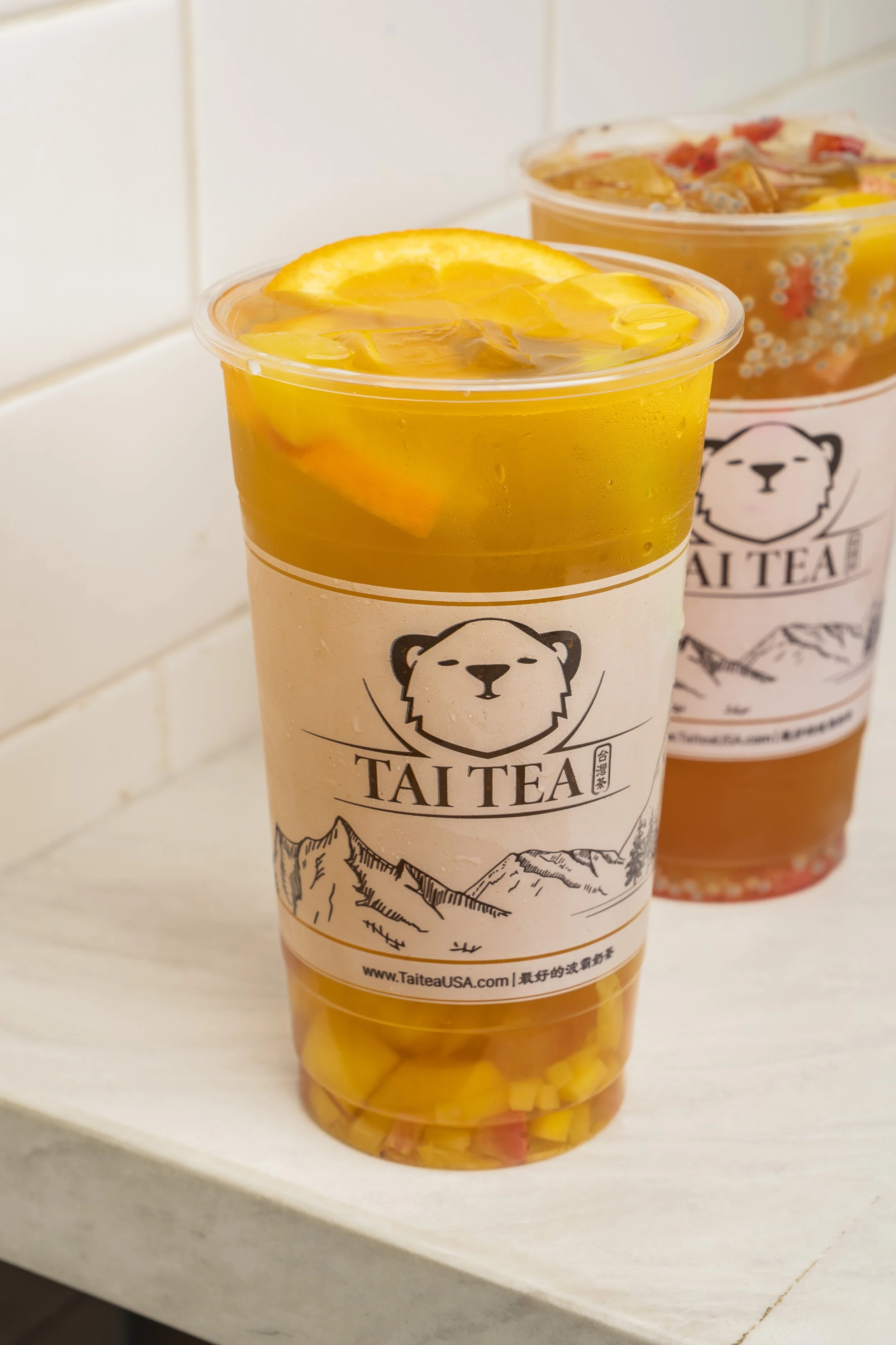 Two large cups of tea with orange slices and colorful jelly cubes at the bottom, labeled 'TAI TEA' with a bear logo and mountain illustration.