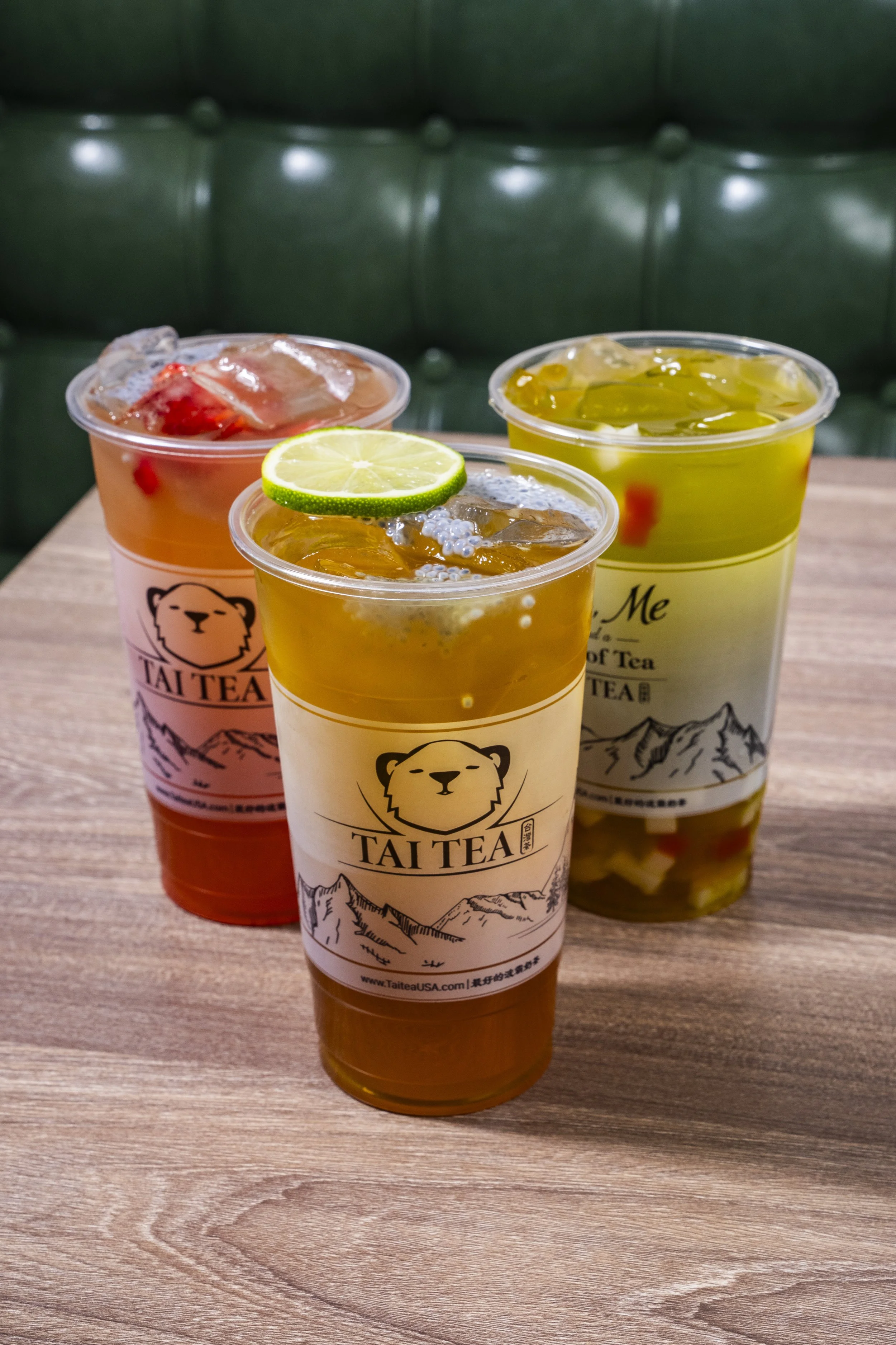 Three cups of bubble tea on a wooden table, with a green padded booth in the background. Each cup has a logo featuring a bear's face and mountains, and the cups contain different colored teas with visible fruit and jelly toppings.