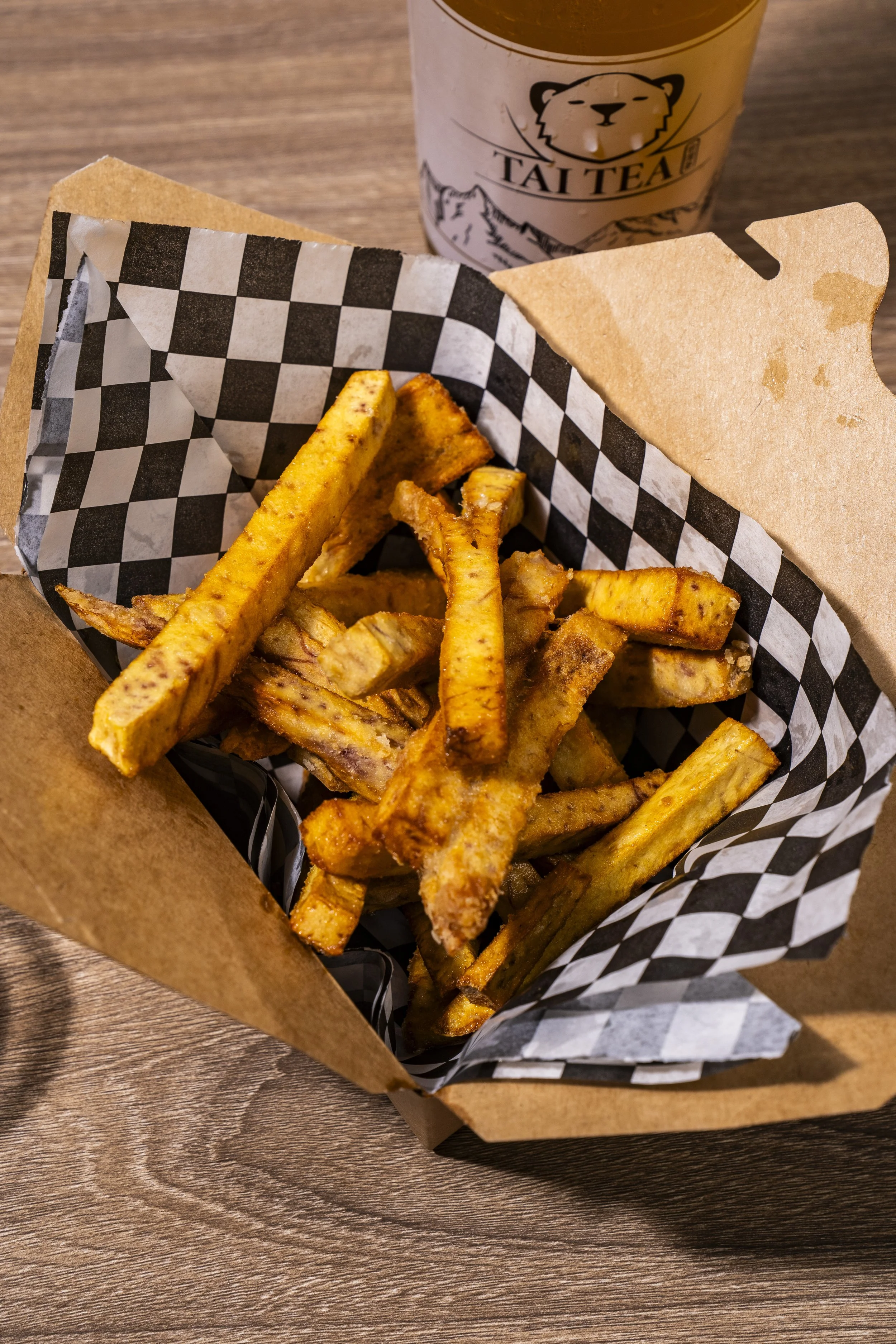Basket of seasoned French fries with a paper lining, placed on a wooden table, with a drink cup labeled 'Tait T' featuring a bear illustration in the background.