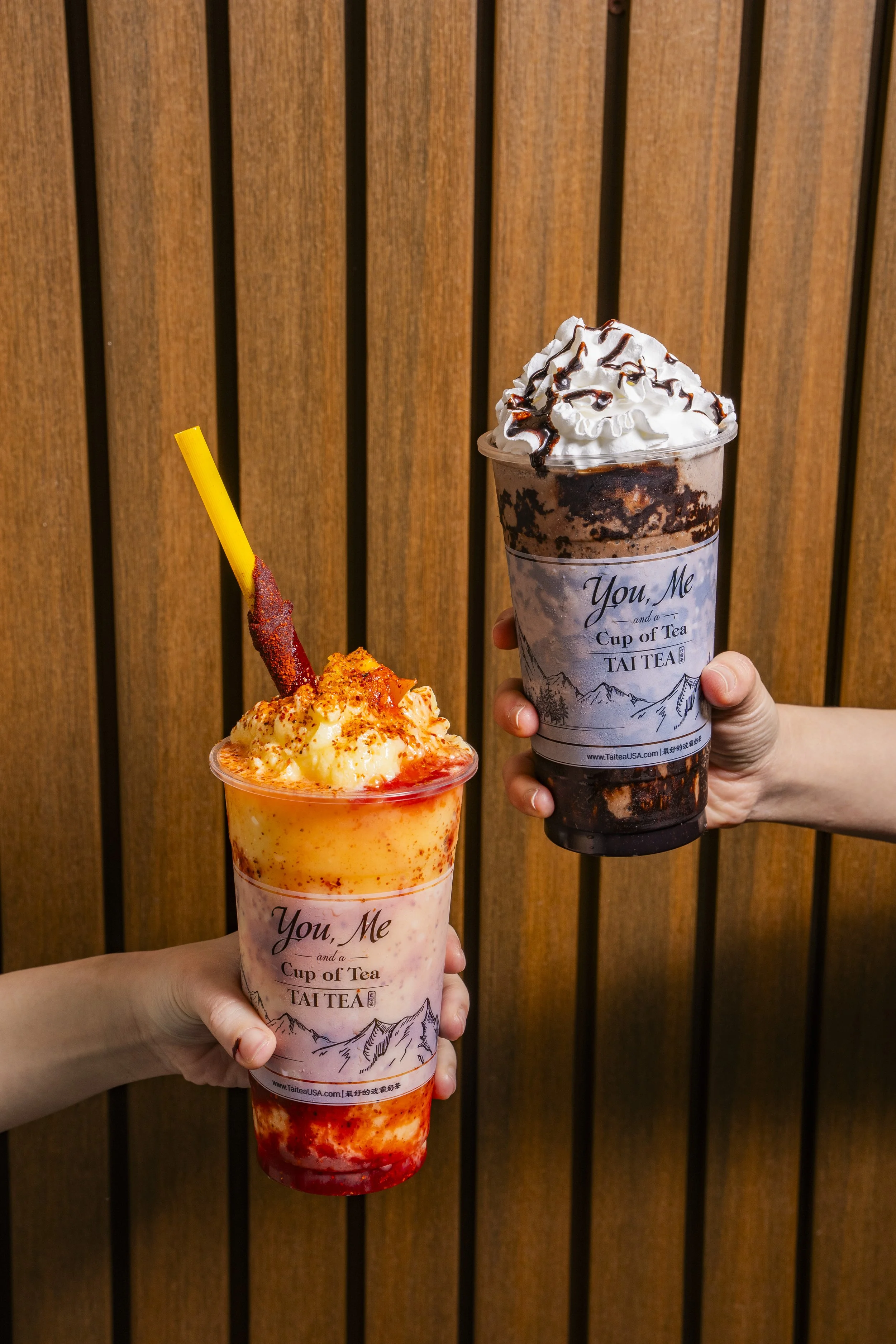 Two hands holding large cups of colorful beverages with whipped cream and decorative toppings against a wooden panel background.