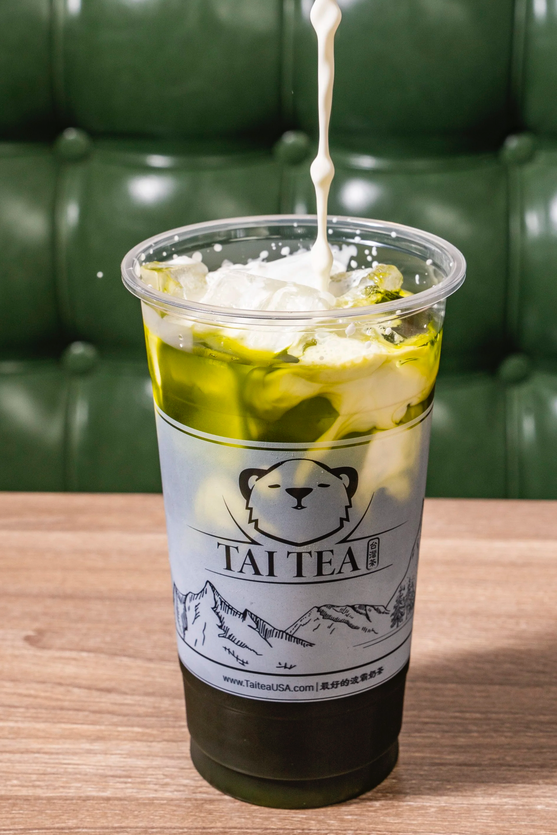 A cold beverage in a transparent cup with ice, cream being poured, and a green and white logo that reads 'TAI TEA' with a bear face and mountain illustration.