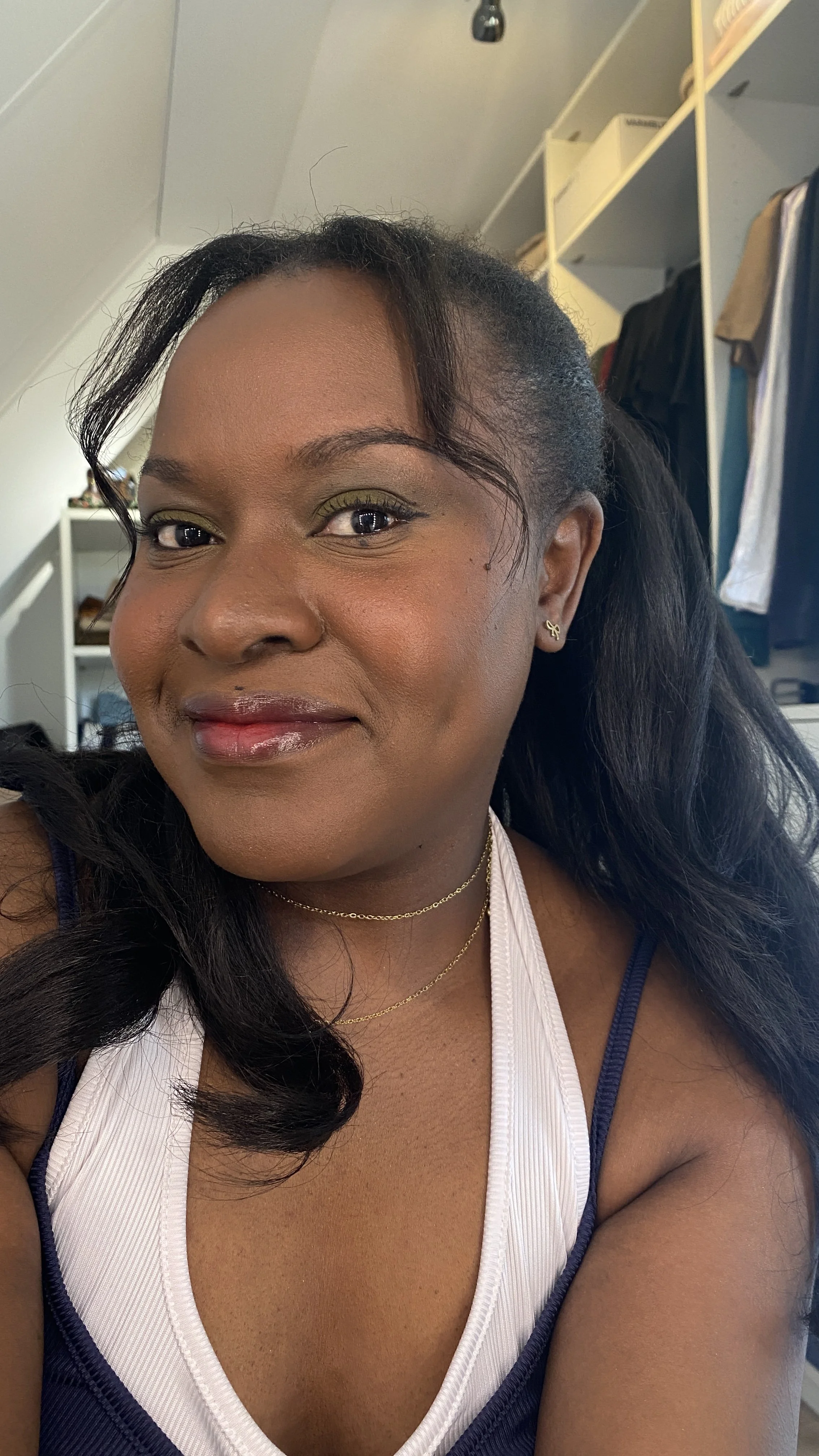 A woman with dark hair, wearing makeup, jewelry, and a navy and white top, smiling in a room with shelves and clothing in the background.