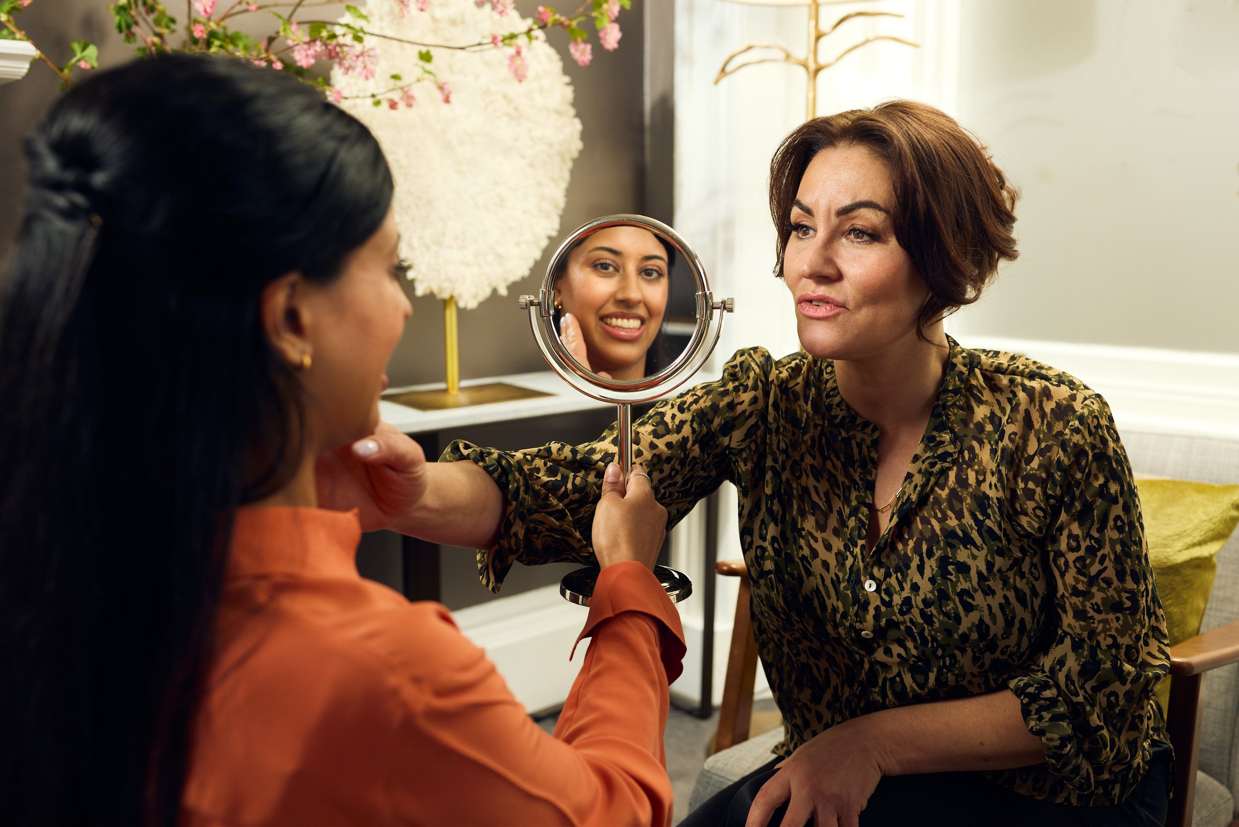 A woman with short brown hair is looking into a small mirror held by another person wearing an orange shirt. The woman appears to be examining her face or makeup. The background shows soft lighting, a floral wall decoration, and a gold coat rack.