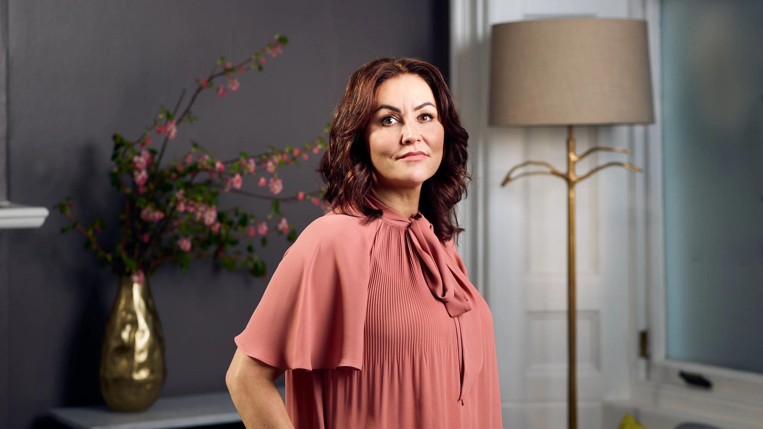 A woman with dark hair and fair skin looking at the camera, wearing a pink pleated blouse with a bow tie at the neckline, standing in a room with a dark wall, a gold vase with pink flowers, and a beige lamp with a gold branch stand in the background.