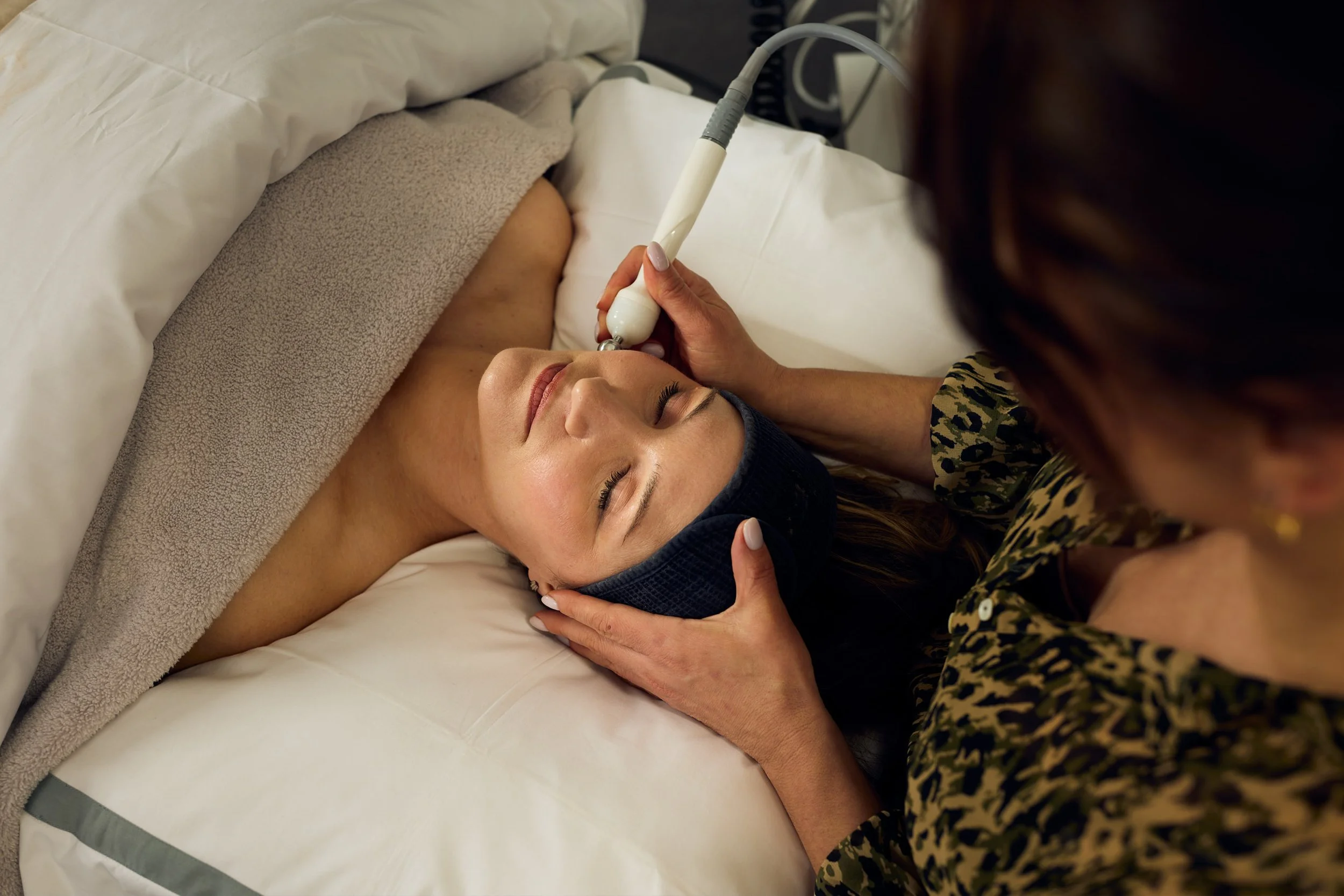 A woman lying on her back with eyes closed, receiving a facial treatment using a handheld device, while a professional gently supports her head in a spa or beauty clinic setting.