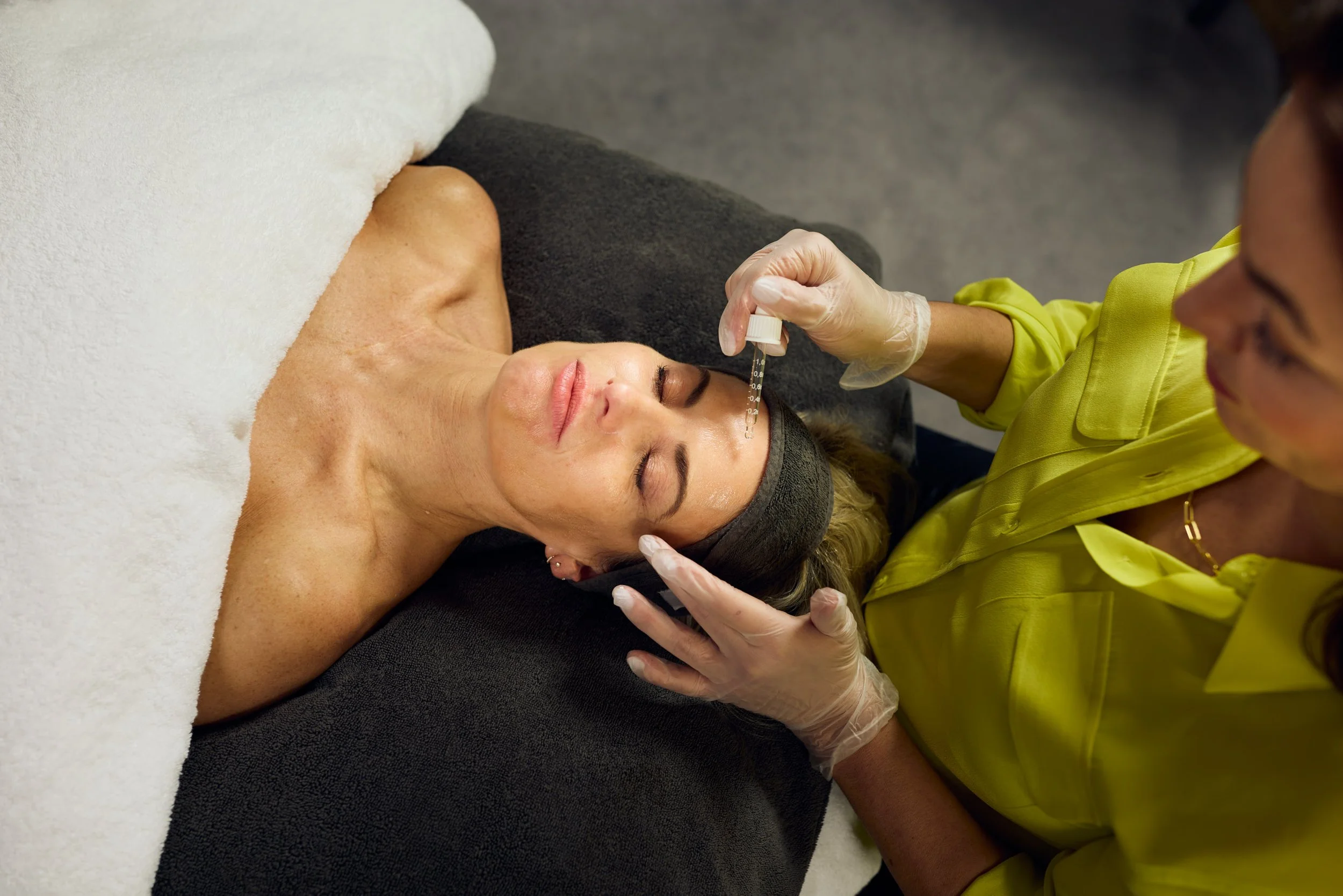 A woman lying on her back with her eyes closed during a cosmetic procedure, receiving a facial treatment with a small dropper or pipette held by a person in a yellow shirt wearing gloves.