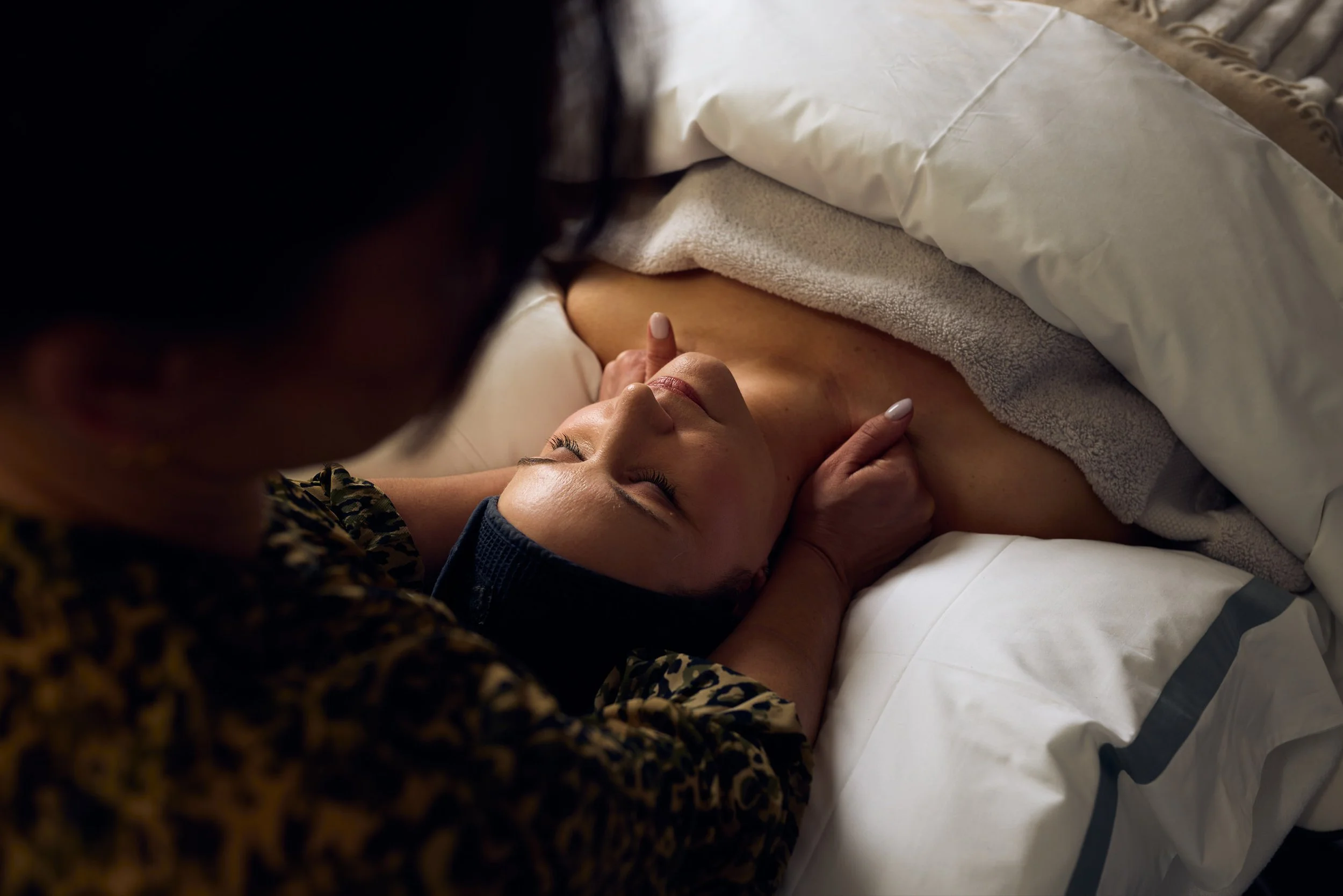 A woman lying on a bed, receiving a massage from a person leaning over her, with a towel covering her chest, and pillows and blankets on the bed.