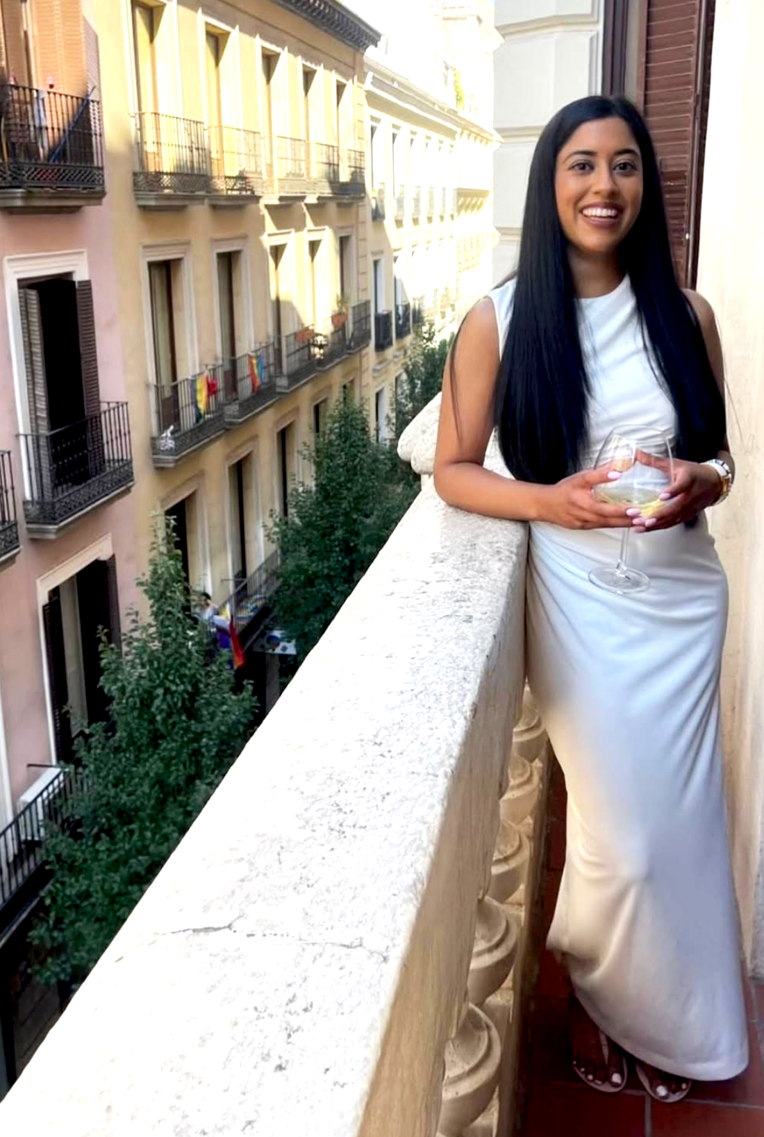 A woman with long black hair smiling and holding a wine glass on a balcony overlooking a European city street with trees and multi-story buildings with balconies.