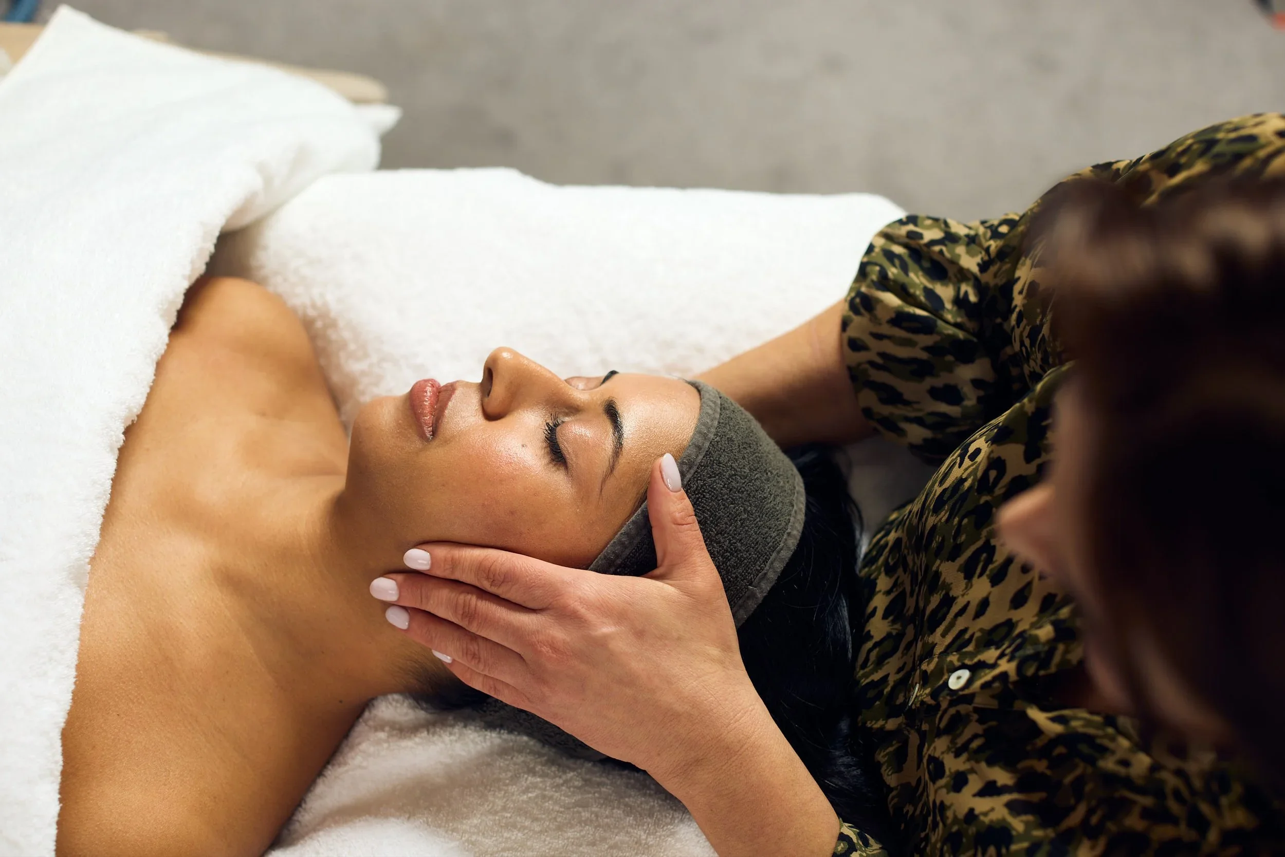 A woman receiving a facial massage from a therapist in a spa setting, lying on a treatment table with a towel and headband.