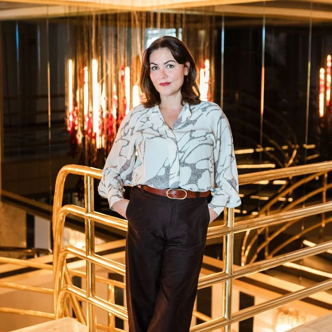 A woman with dark hair dressed in a patterned blouse and dark trousers standing on a gold-colored staircase railing, with a background of modern interior decor featuring warm lighting and vertical light fixtures.