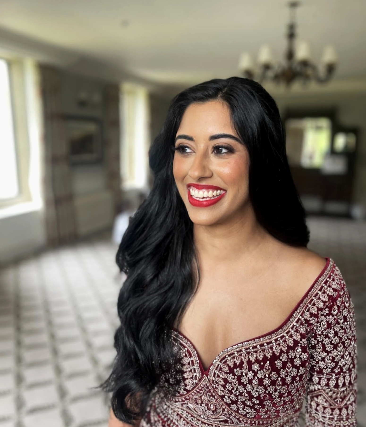 A woman with long black hair, bright red lipstick, wearing a traditional maroon embroidered outfit, smiling in a well-lit room with chandeliers and windows.