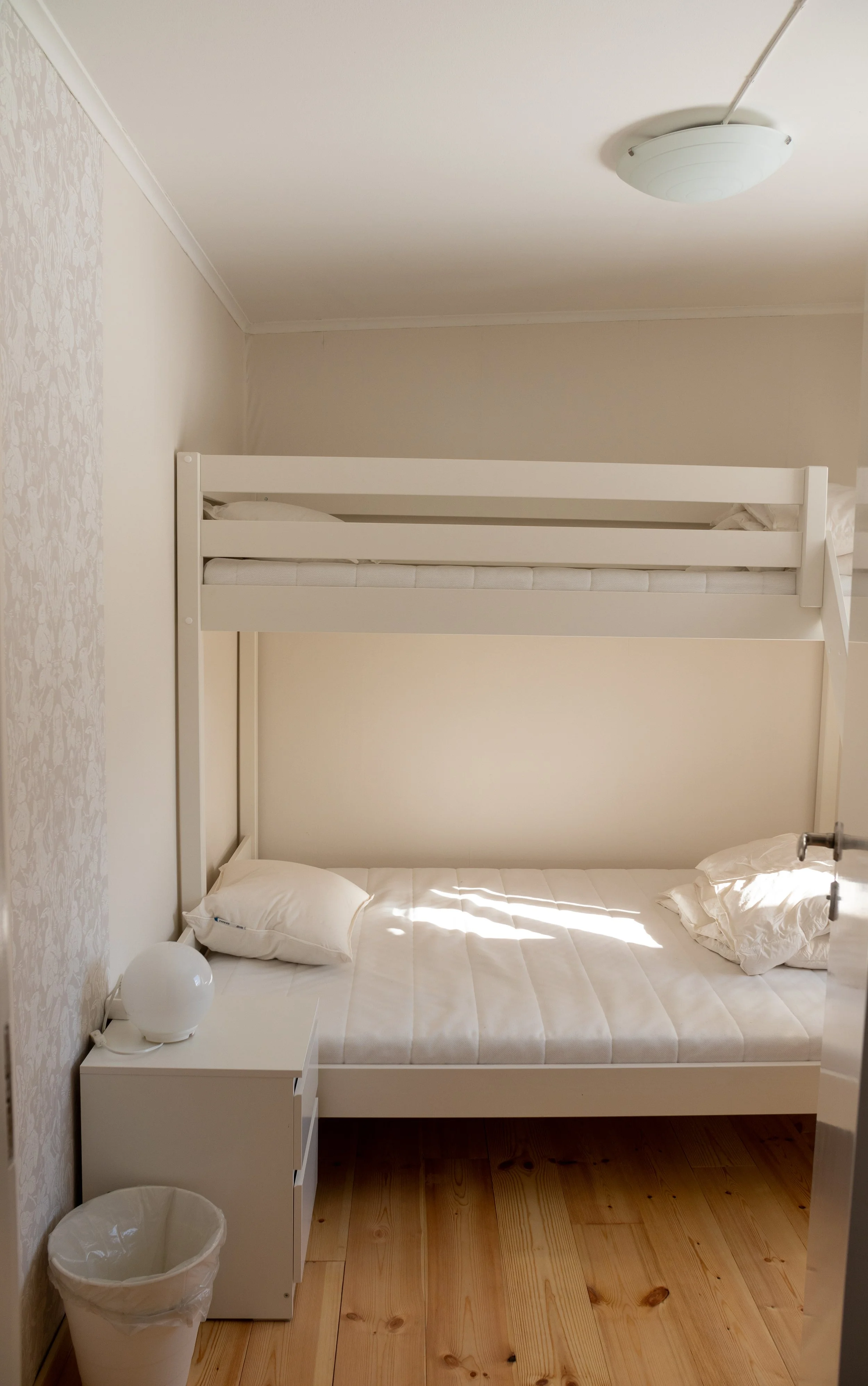 Ein Schlafzimmer mit einem weißen Etagenbett, einem Nachttisch mit einer Schreibtischlampe, zwei weißen Kissen, einem Wanddekor an der linken Wand, und einem Holzboden.