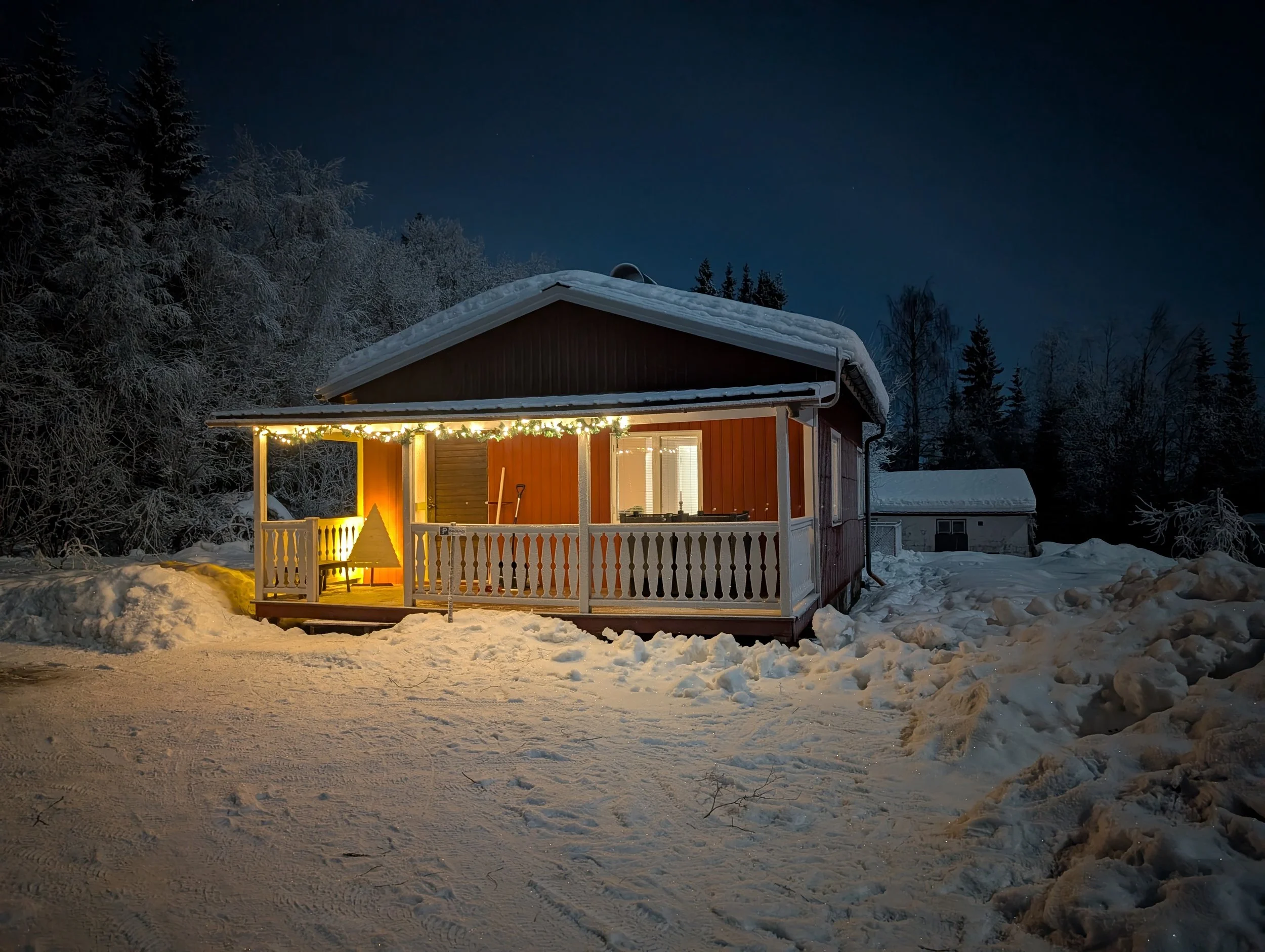 Ein kleines, rot gestrichene Haus mit beleuchteter Veranda steht im Schneefall bei Nacht, umgeben von Schnee bedeckten Bäumen.