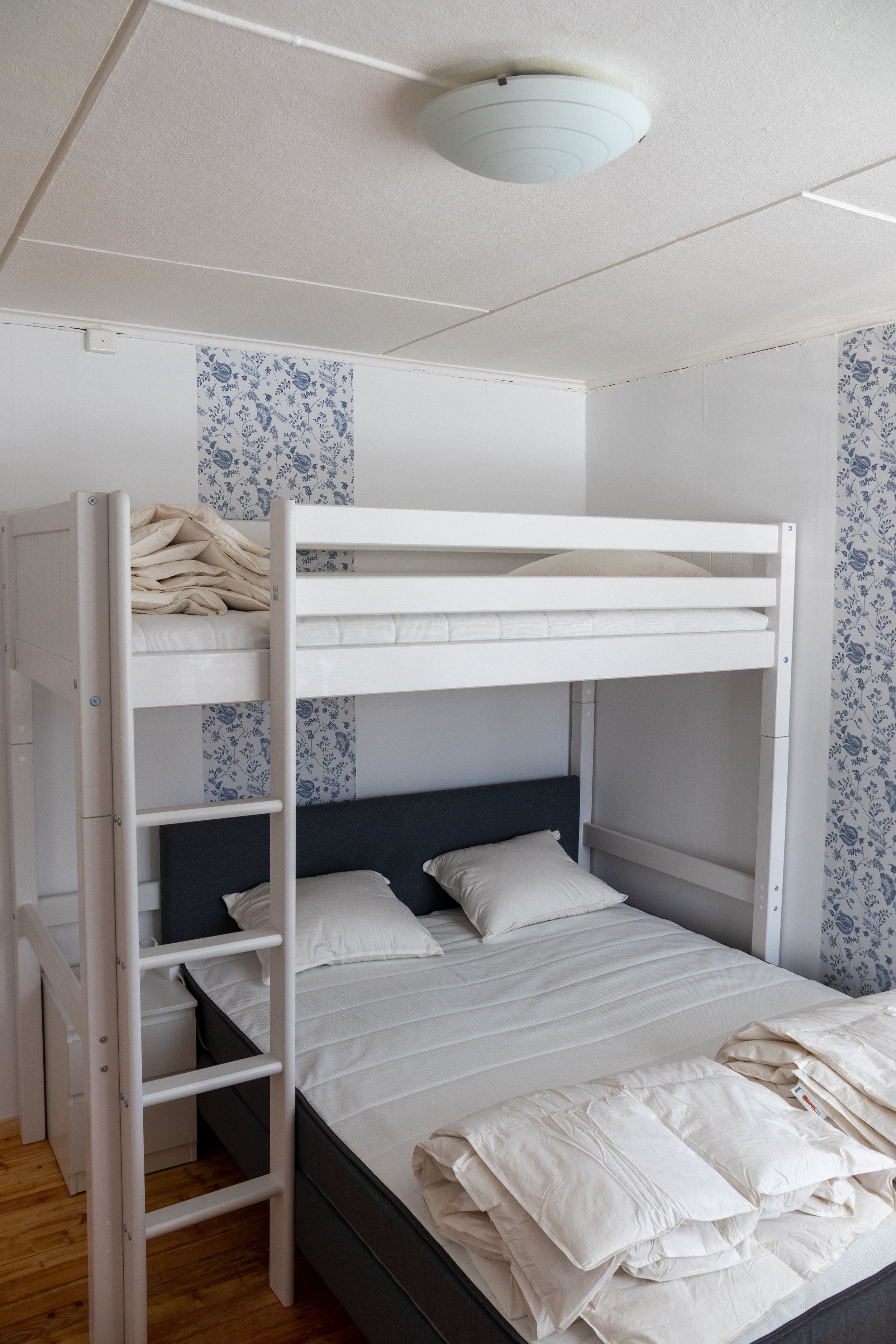 Ein Schlafzimmer mit einem Doppelbett und einem Hochbett, das aus weißem Holz besteht. Das Doppelbett hat weiße Bettwäsche und Kissen, während die Bettwäsche auf dem unteren Bett offen ist.