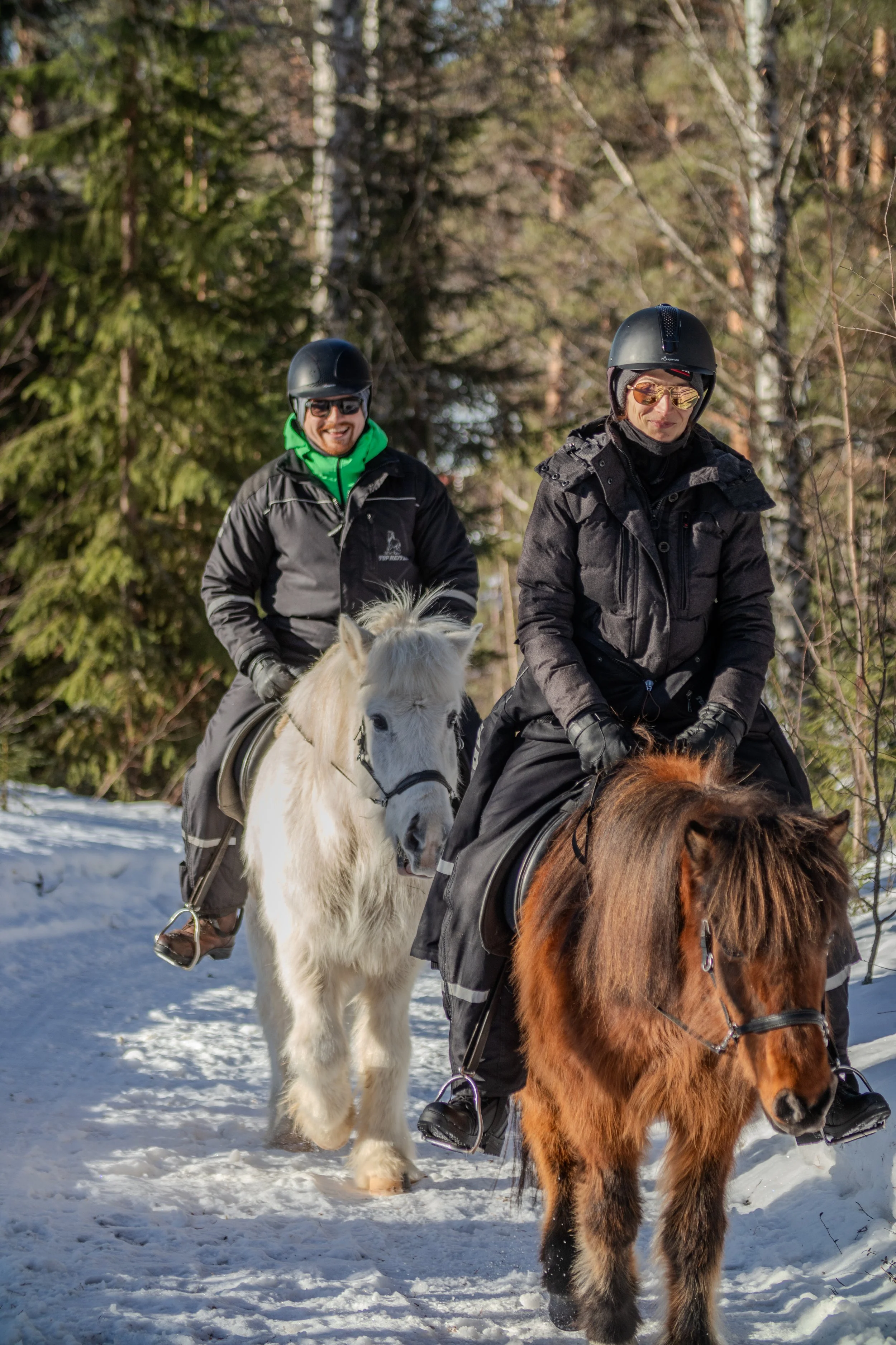 Zwei Menschen reiten auf Pferden durch den Schnee in einem bewaldeten Gebiet. Beide tragen Helme und Winterkleidung.