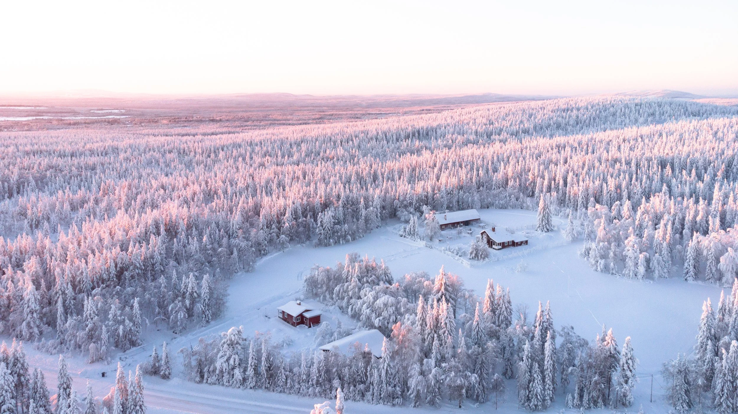 Eine verschneite Winterlandschaft mit mehreren roten Holzhäusern, umgeben von gefrorenen Bäumen, mit einem pastelfarbenen Himmel im Hintergrund.